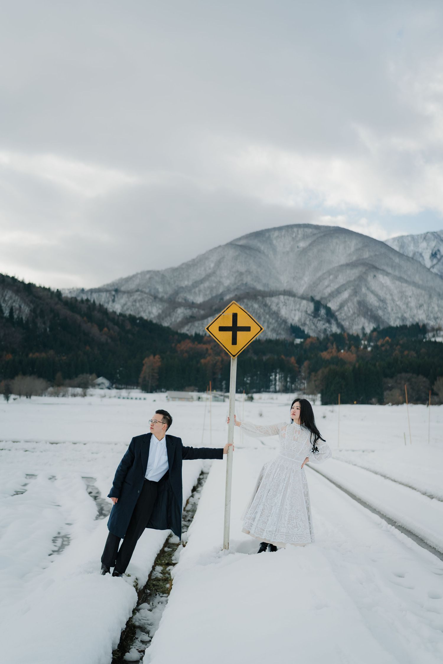 A couple poses playfully by a railroad crossing sign in a snowy mountain landscape.
