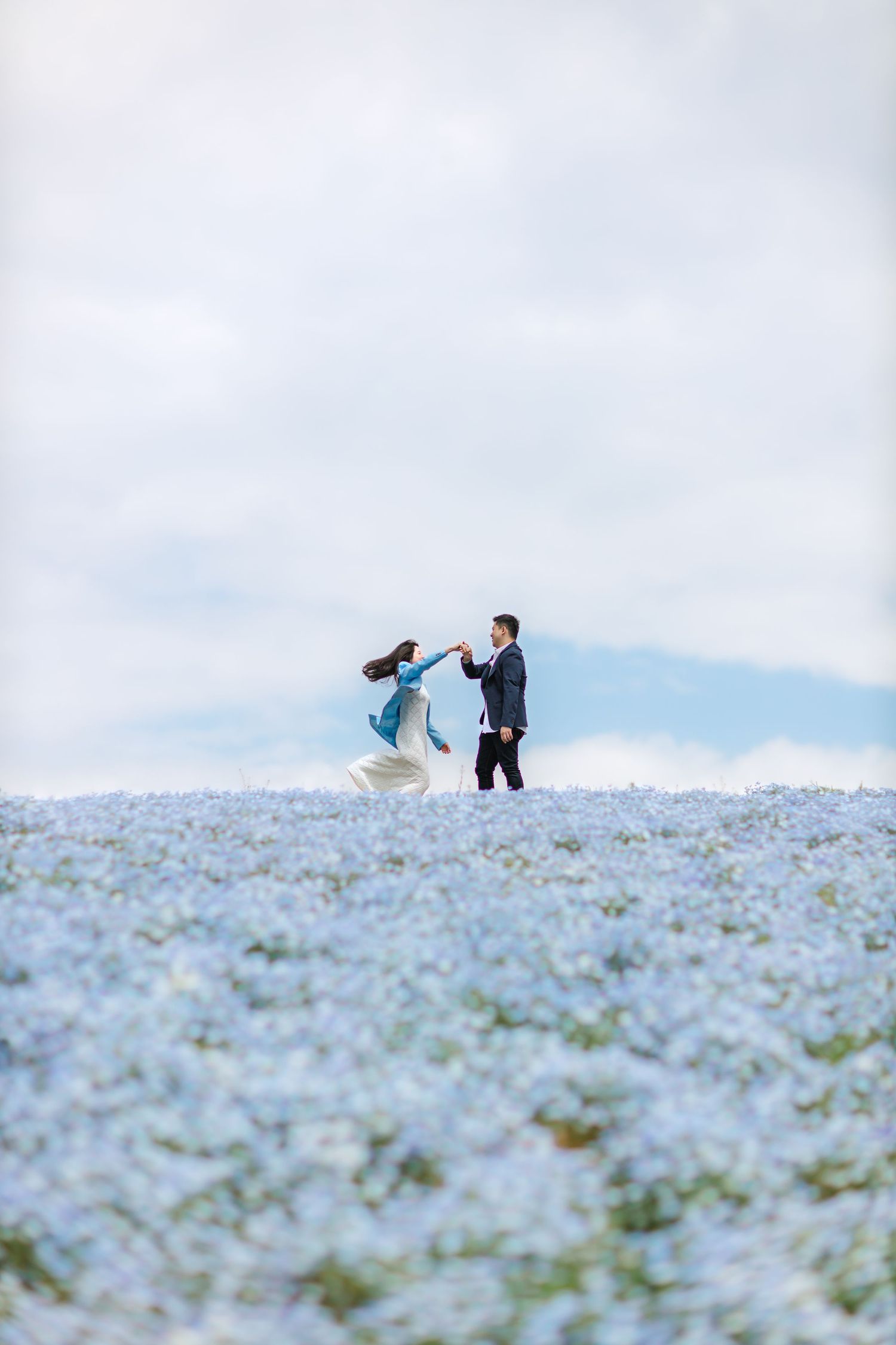 Couple standing in a field of light blue flowers under cloudy skies.