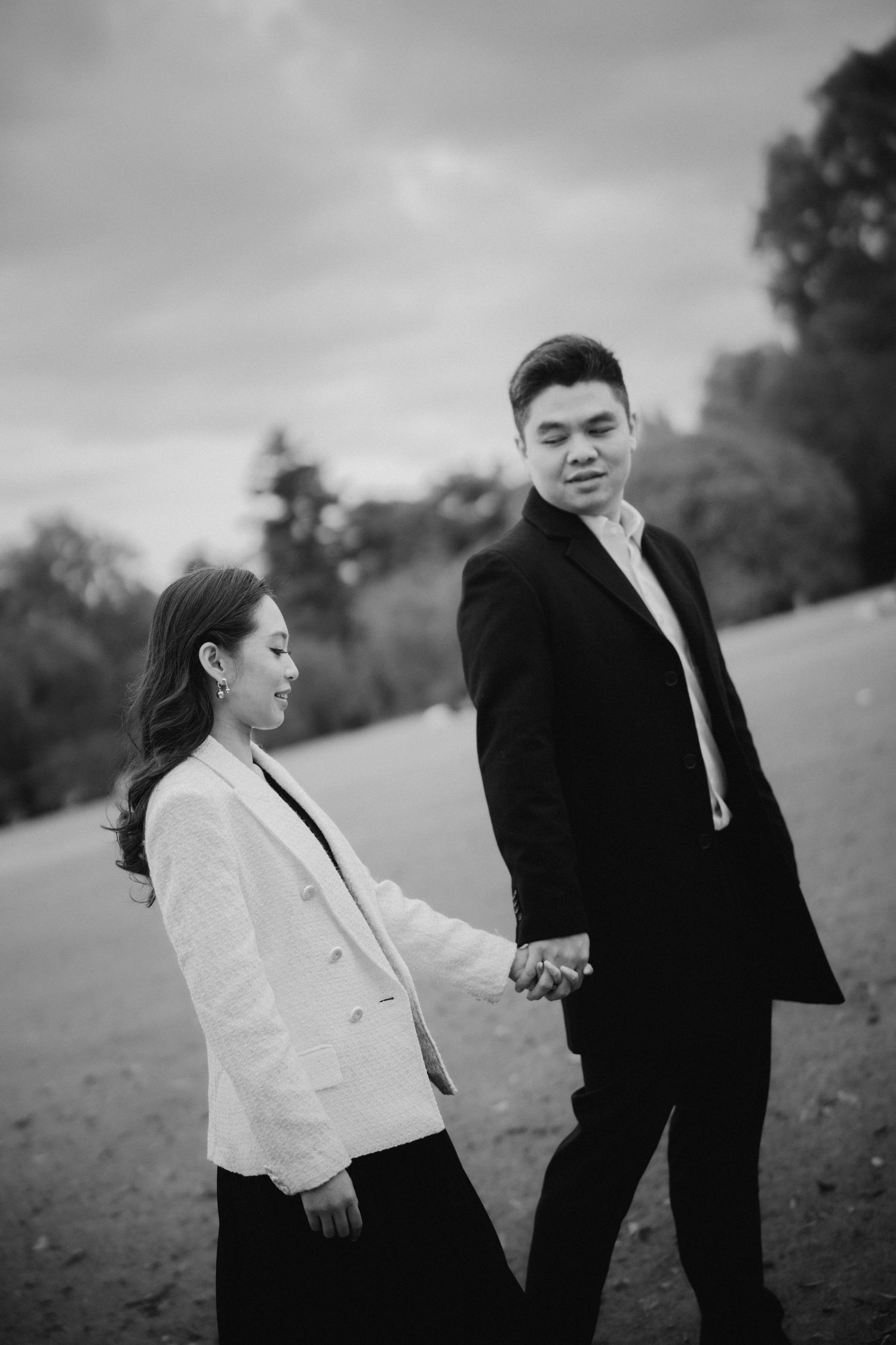 Couple in formal attire holds hands while standing in a misty outdoor setting.