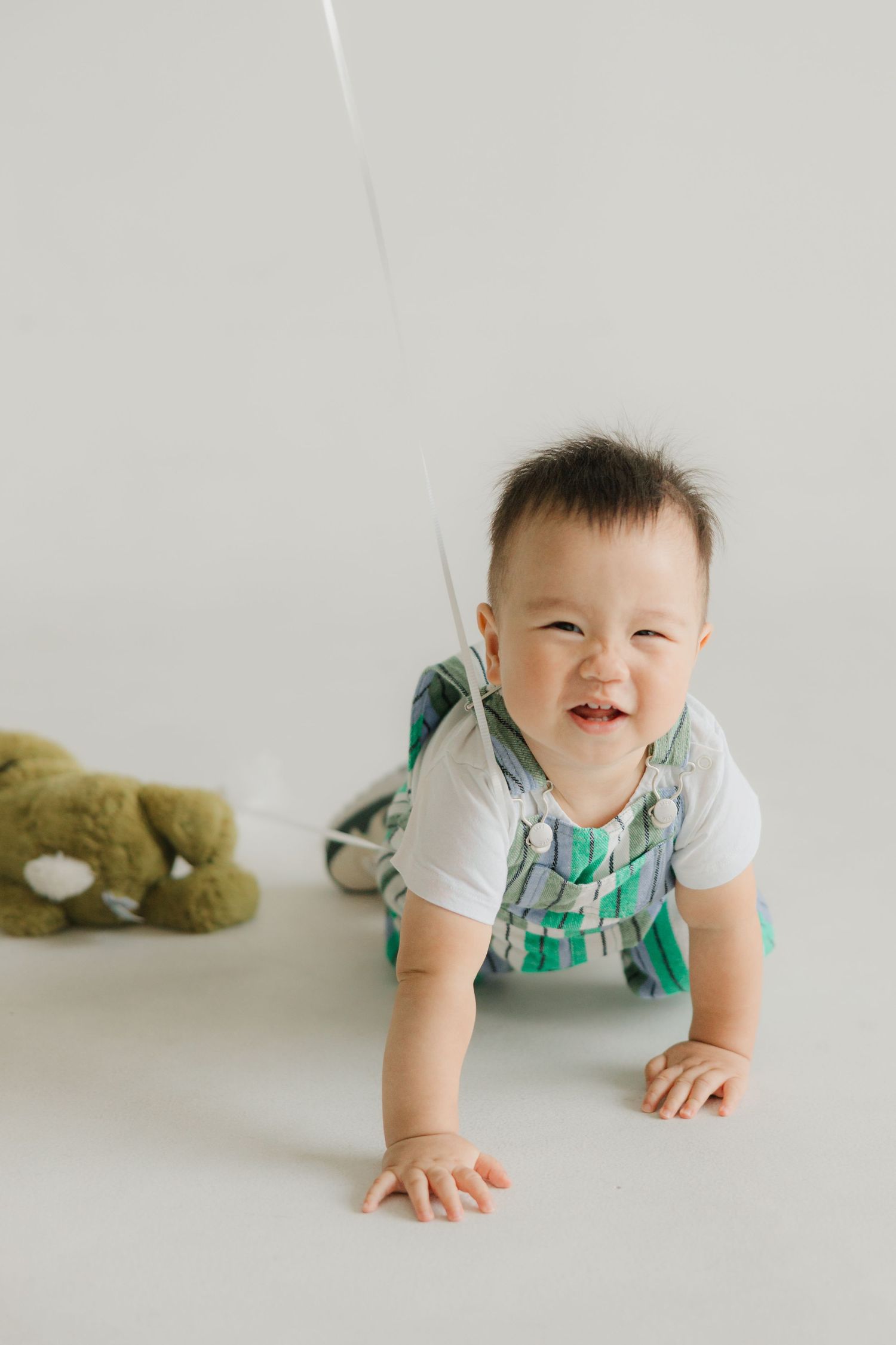 Baby in overalls crawls on white background with stuffed toy nearby.