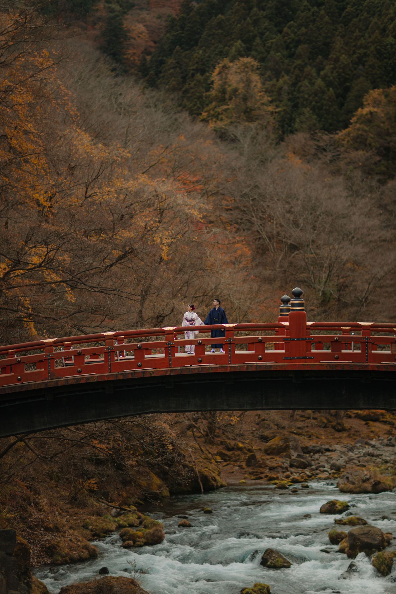 Couple on a red bridge over rushing waters with autumn foliage in the background.