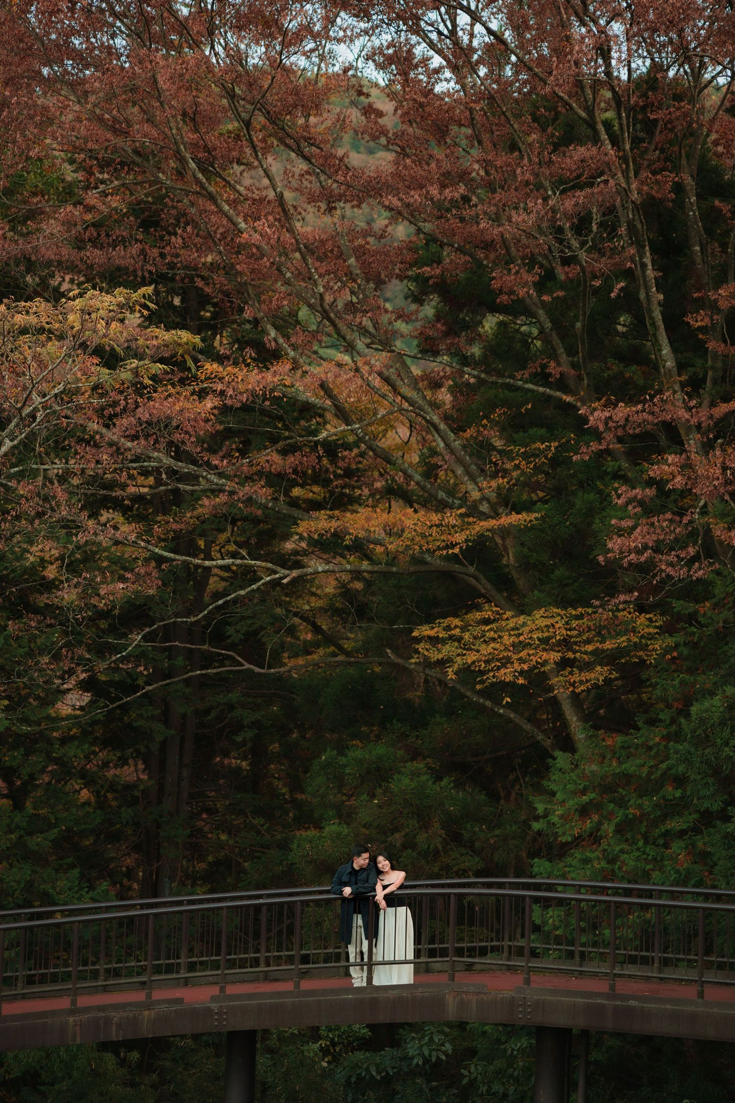 Couple standing on wooden bridge surrounded by autumn foliage in a Japanese garden.