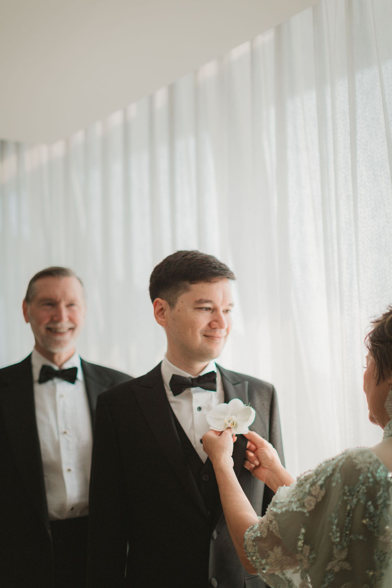 A formal wedding preparation scene with groomsmen in black tuxedos and bow ties against a white curtained backdrop.