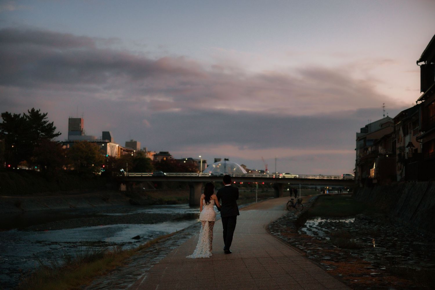 A couple walking along a riverside path at dusk with city lights twinkling in the background.