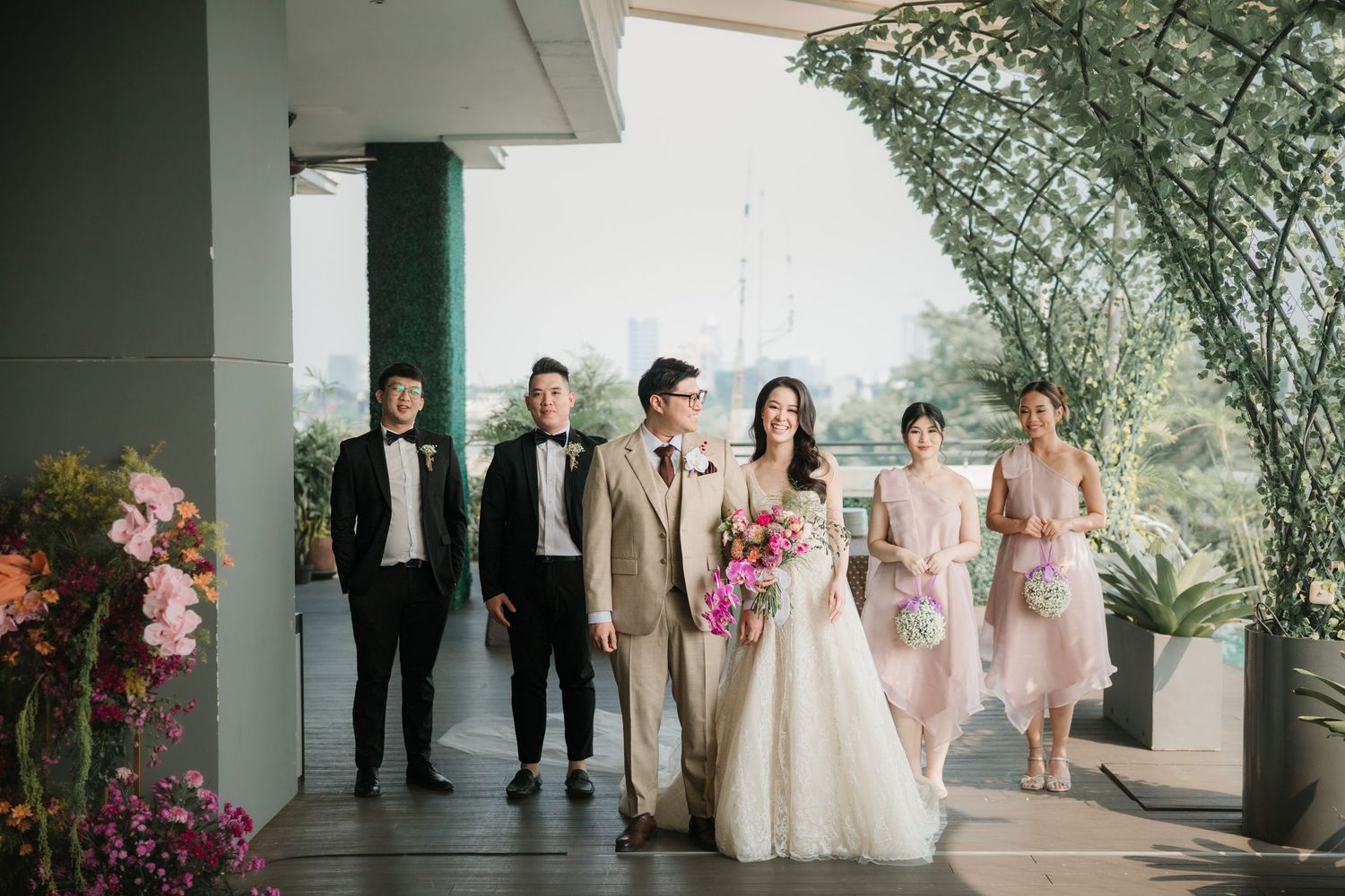 A wedding party poses outdoors with bridesmaids in pink dresses and groomsmen in formal suits.