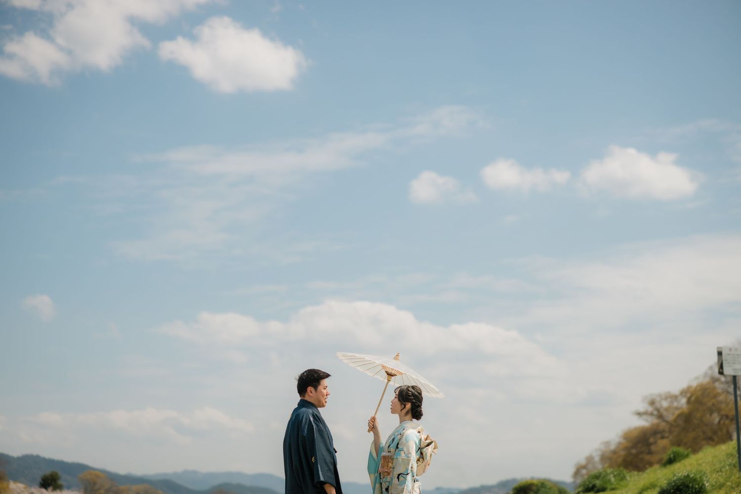 Couple standing on hillside with mountains and blue sky in background.