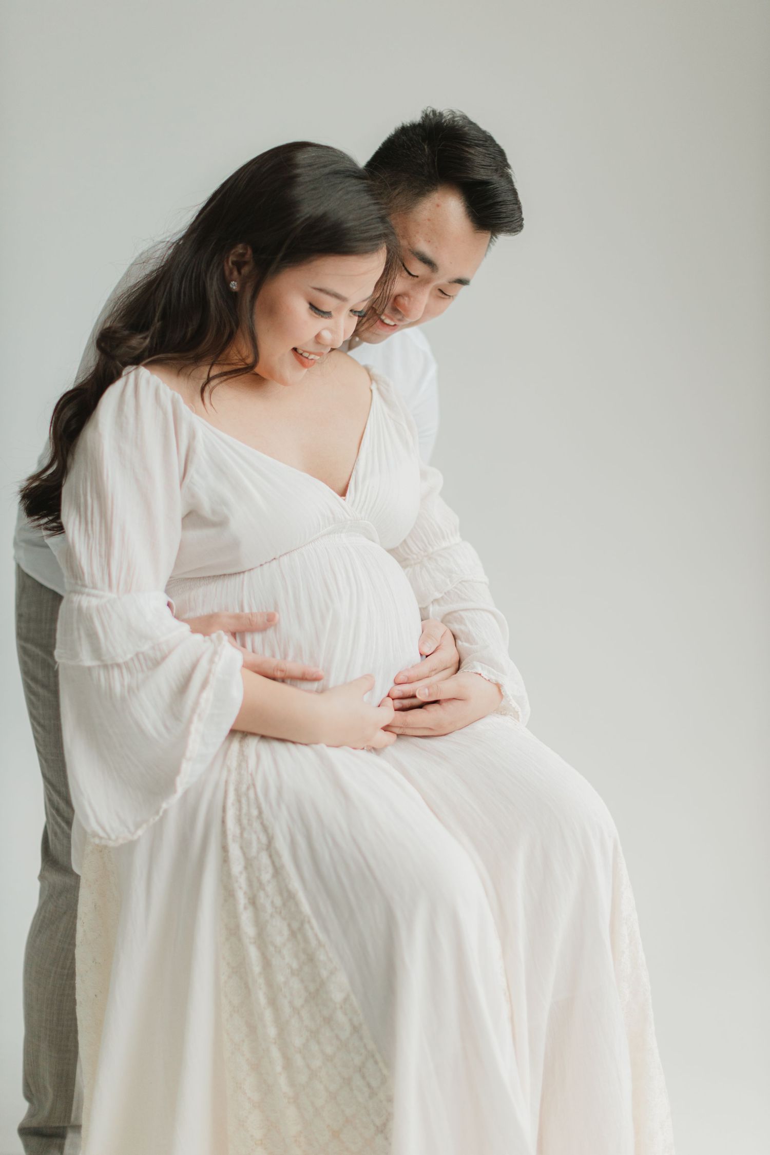 Intimate maternity photo of couple in white clothing embracing against neutral background.