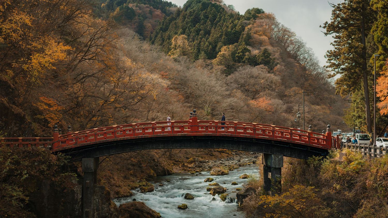 Traditional red Japanese bridge spans a rocky stream amid autumn foliage in a serene mountain landscape.