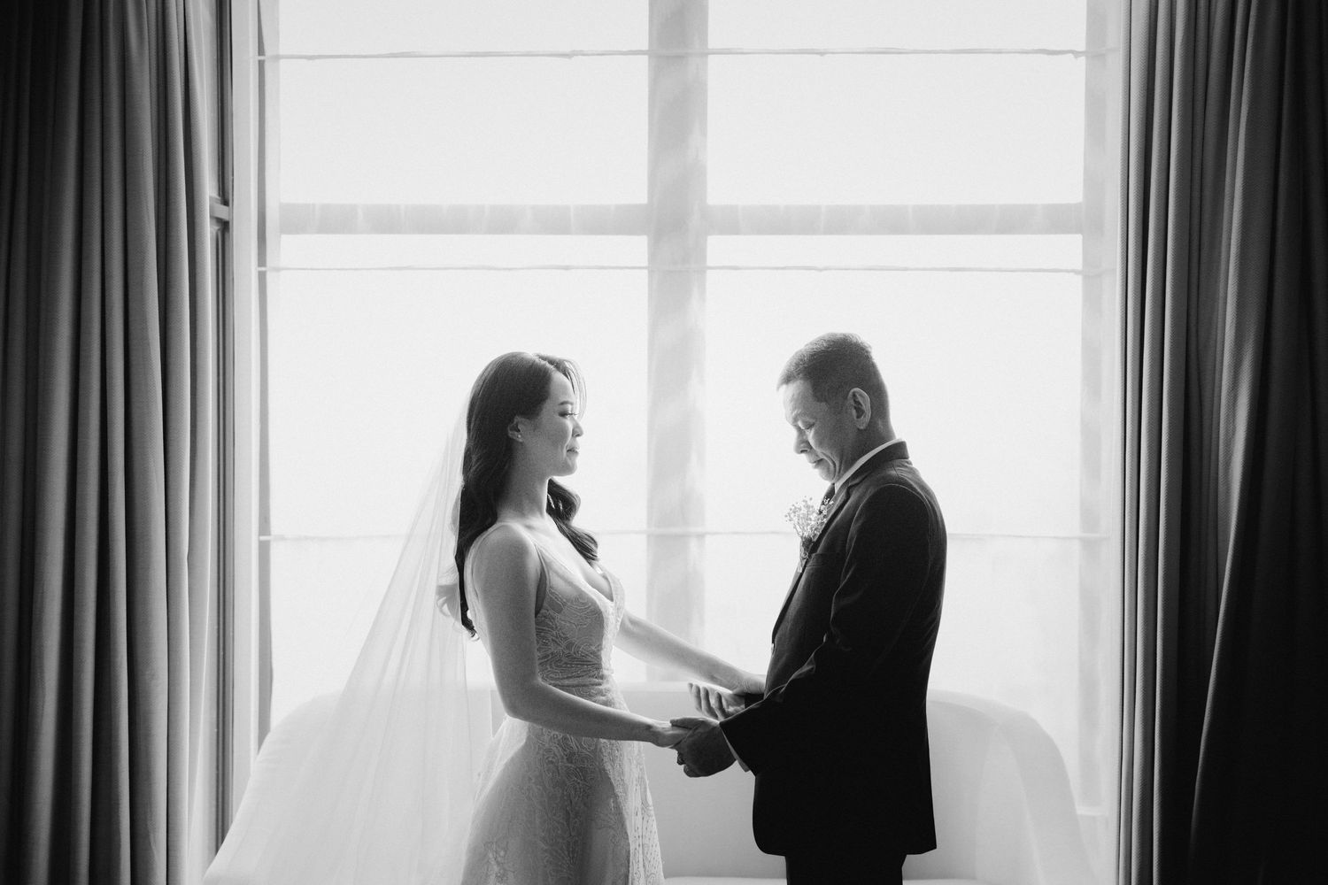 A black and white wedding photo of a couple sharing an intimate moment by a window.