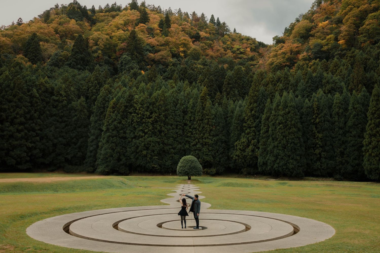 Couple standing in center of circular maze pattern in a garden surrounded by evergreen trees.