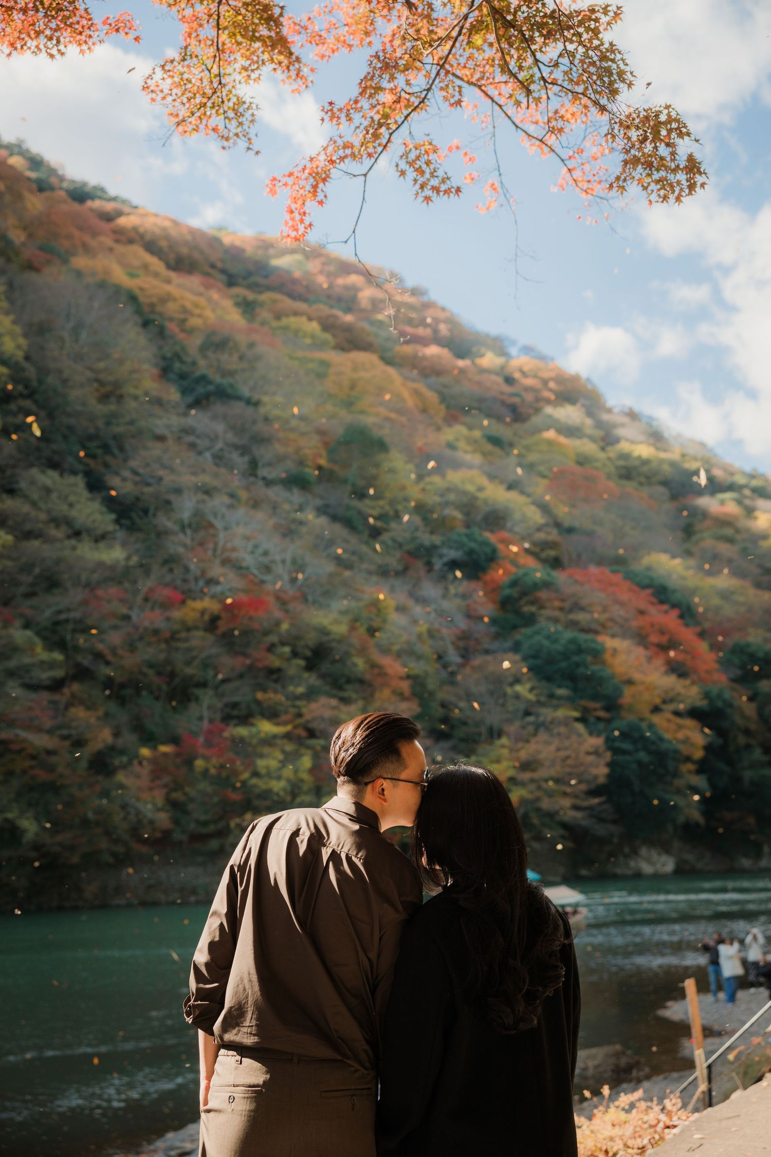 A couple embraces under autumn foliage beside a tranquil lake.