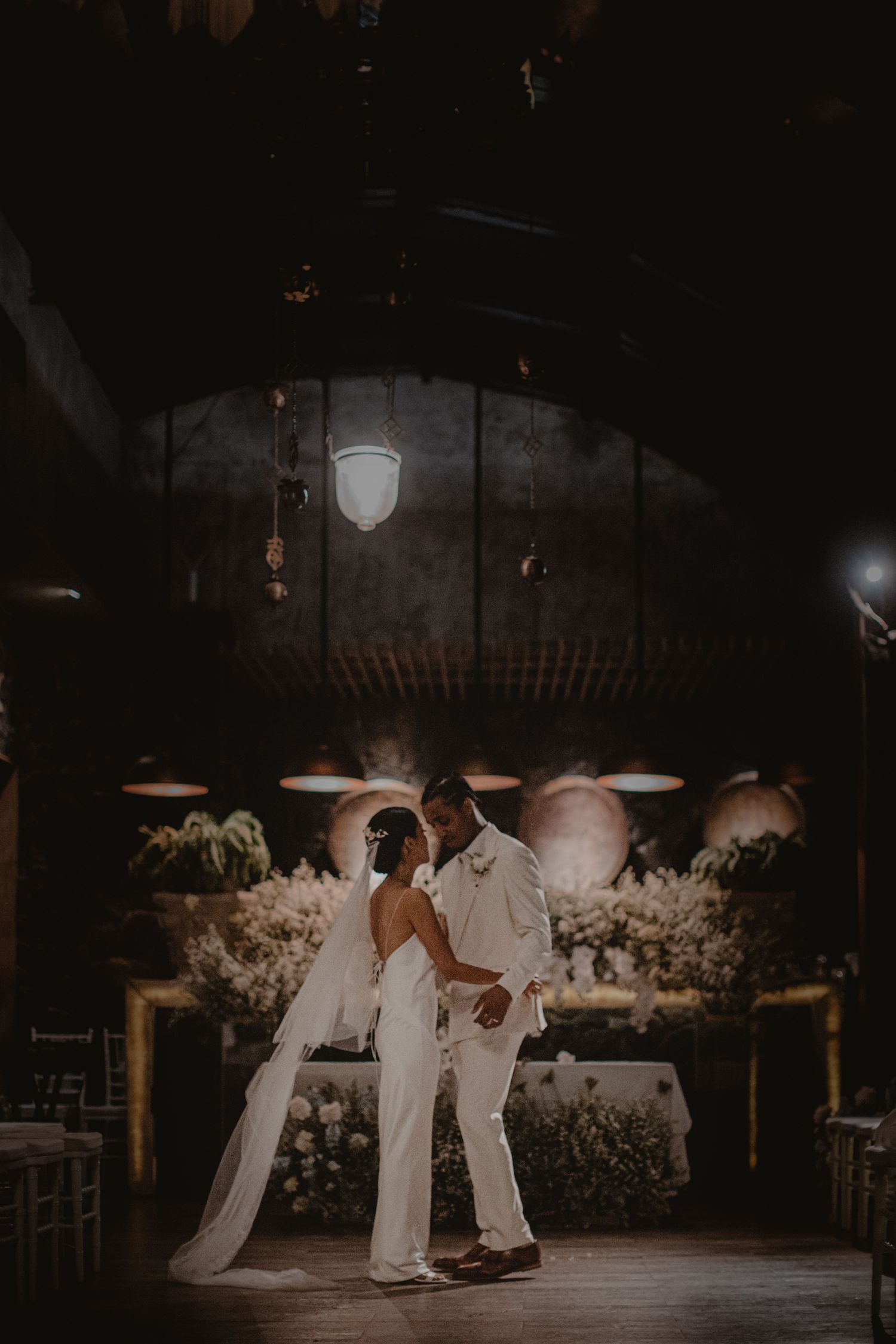 A couple in white outfits dancing together under romantic lighting at their wedding reception.