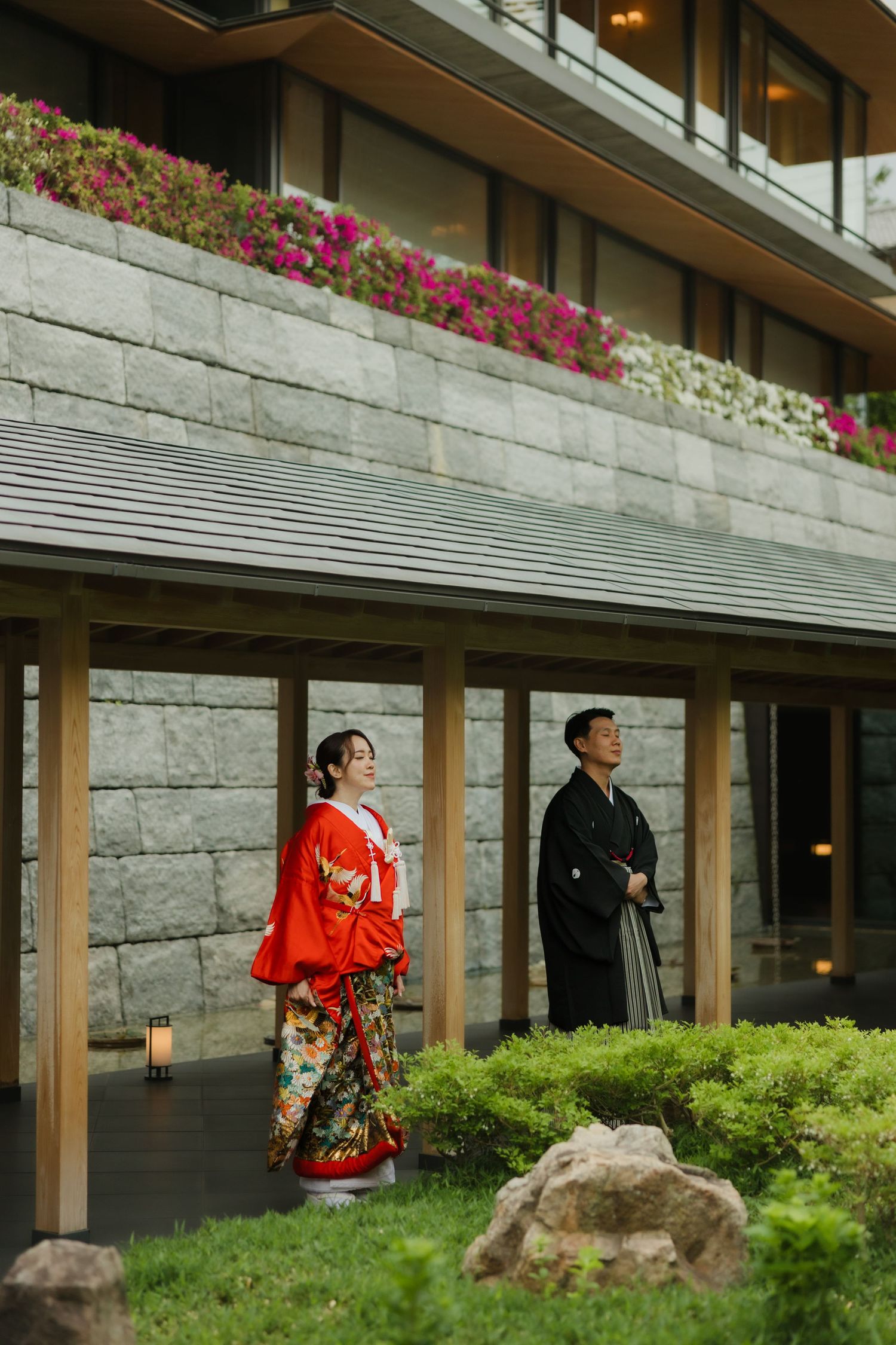Two figures in traditional Japanese attire stand under a stone building adorned with pink and white flowers.