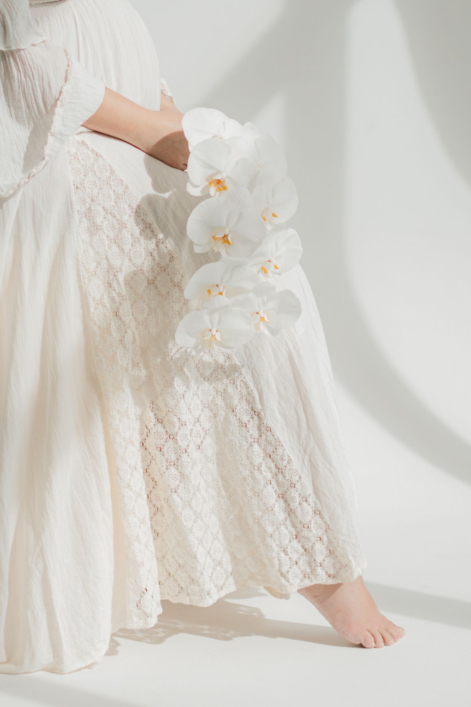Close-up of white lace maternity dress with delicate orchids against light background.