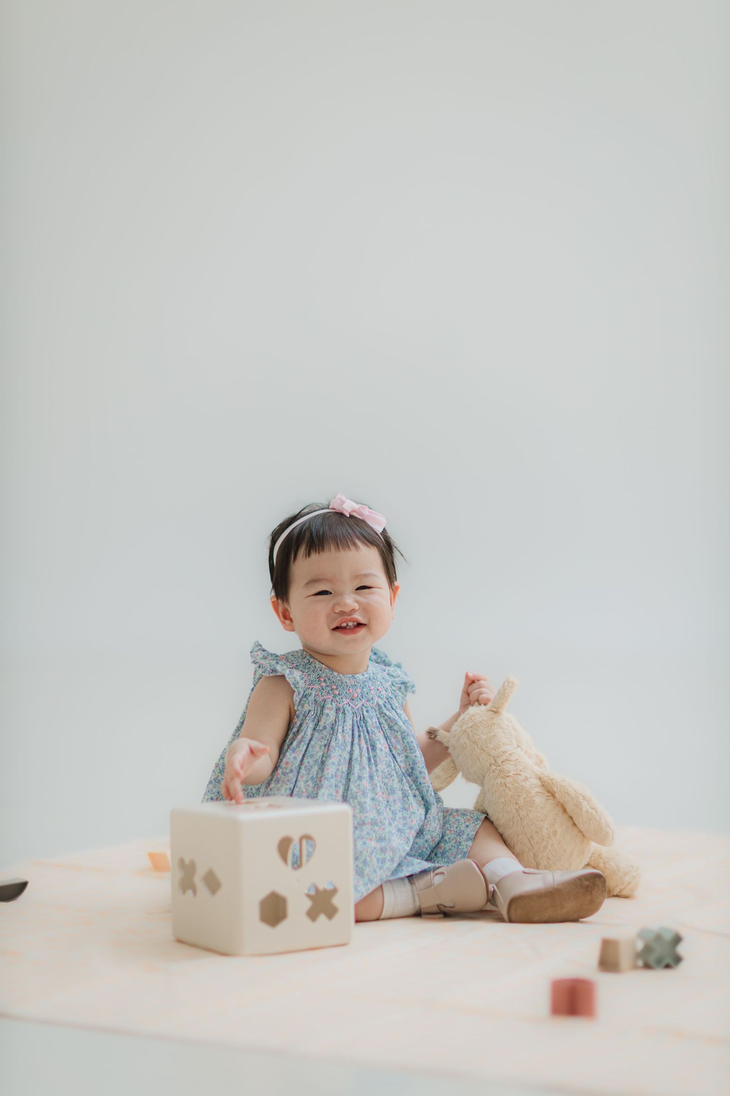 A baby in a blue dress sits with toys and a stuffed animal against a white background.