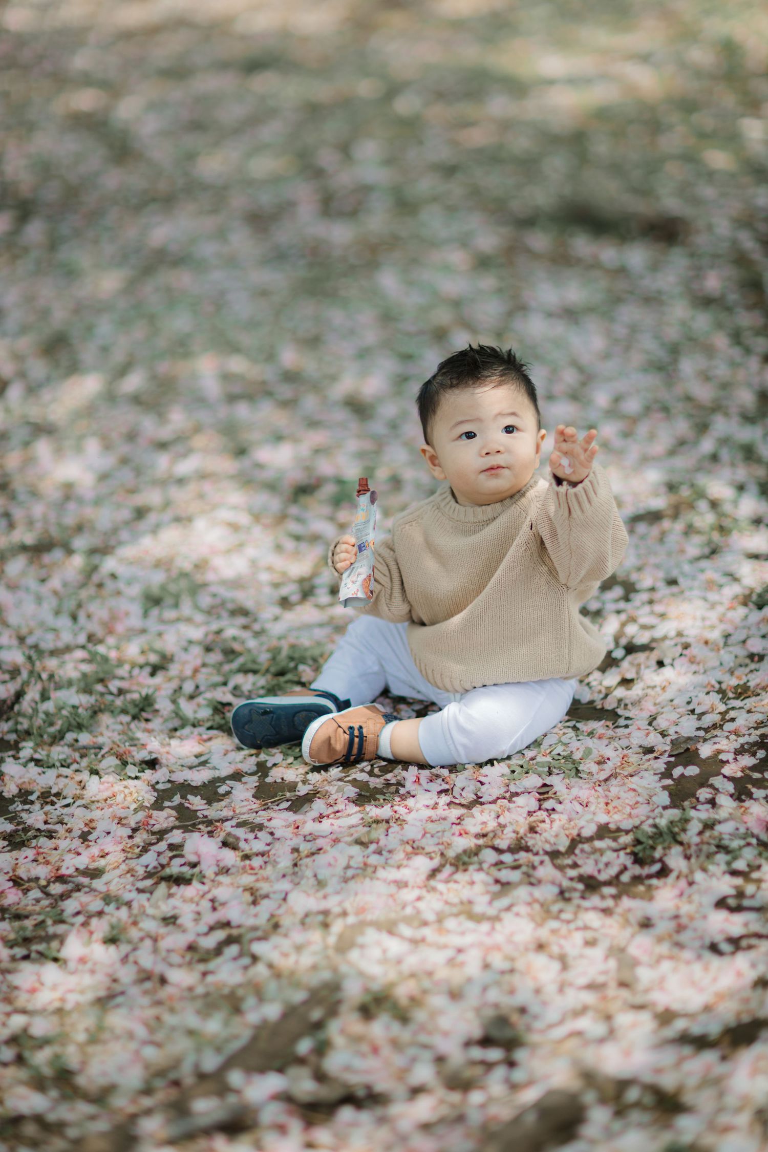Child in beige sweater sits on fallen leaves during autumn.