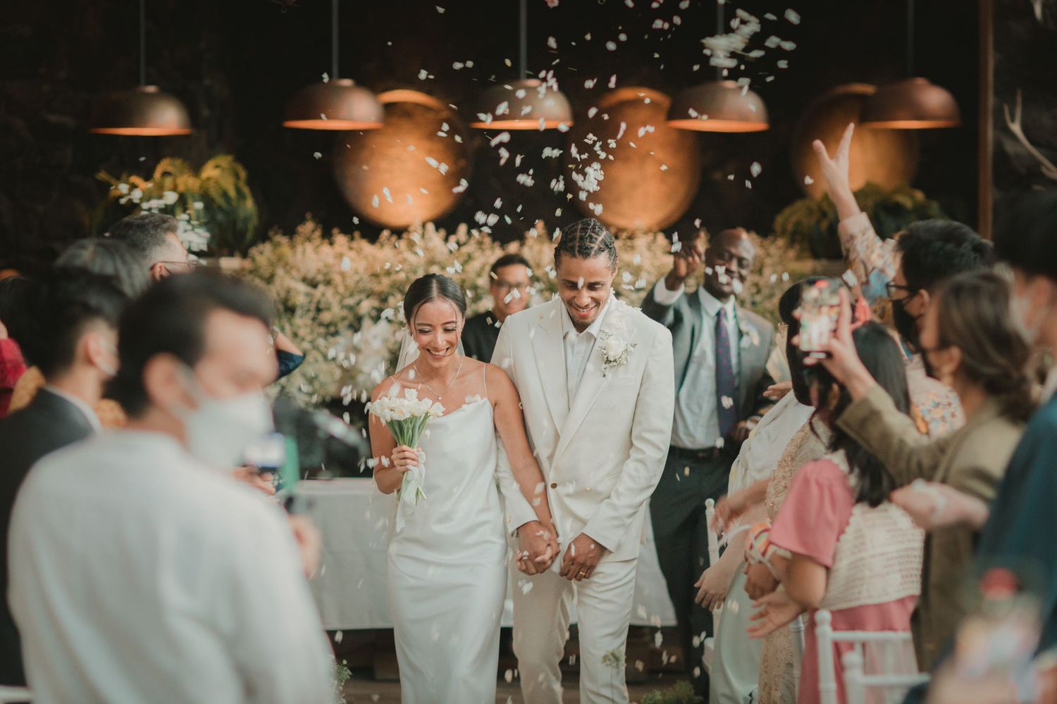 A newlywed couple in white attire walking through a venue with guests throwing confetti.
