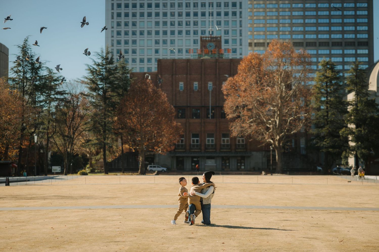 Small figures standing in an urban park with tall buildings and flying birds in the background.