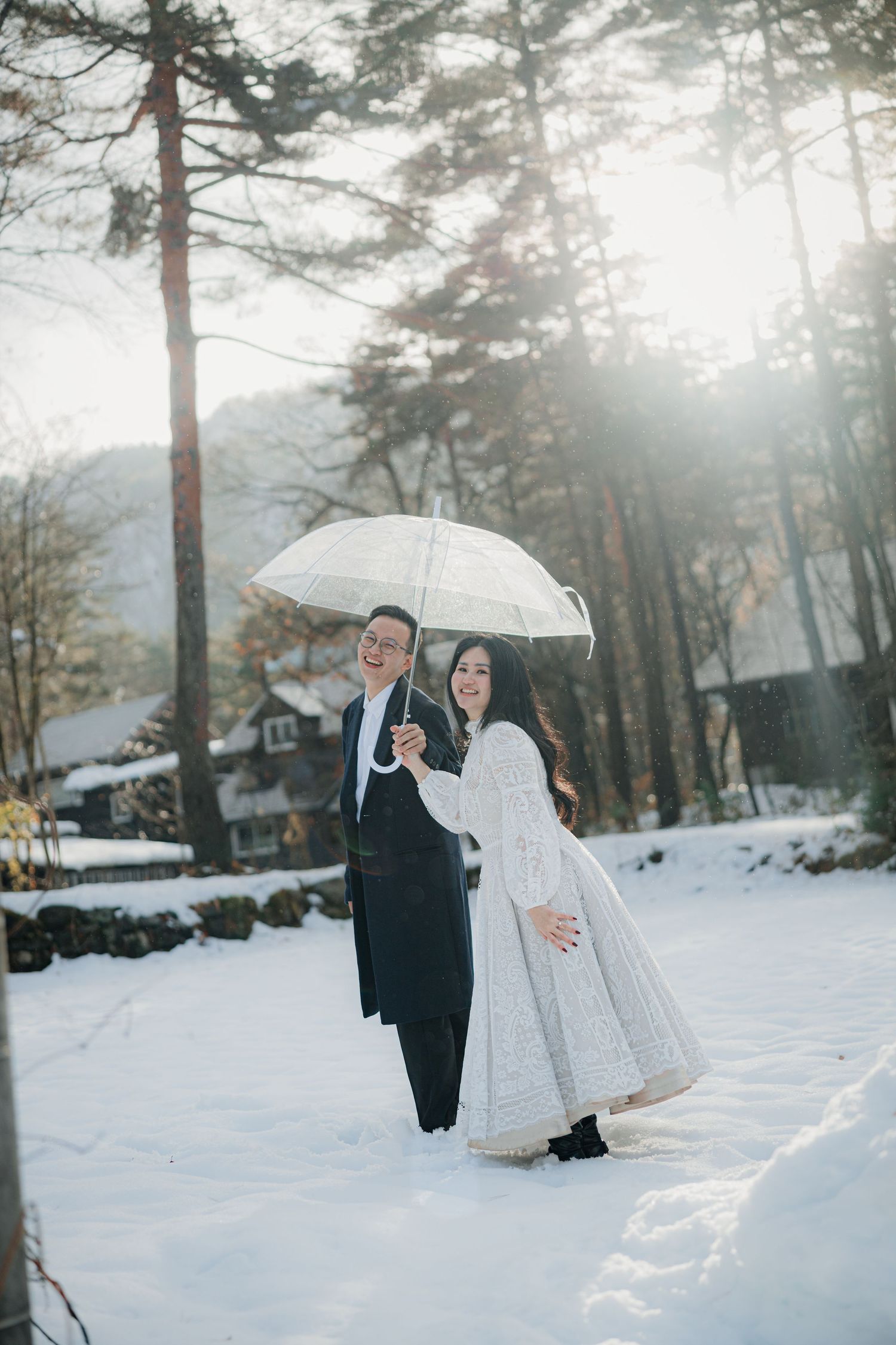 A winter wedding photo with a couple sharing an umbrella in a snowy forest setting.