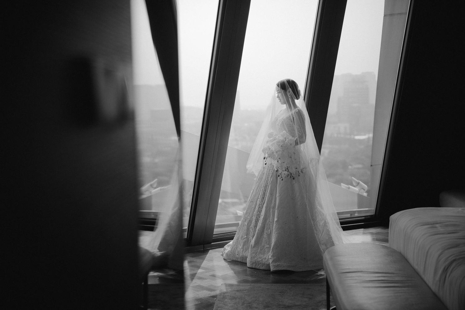 A silhouetted bride in a wedding gown stands by a large window overlooking a cityscape.