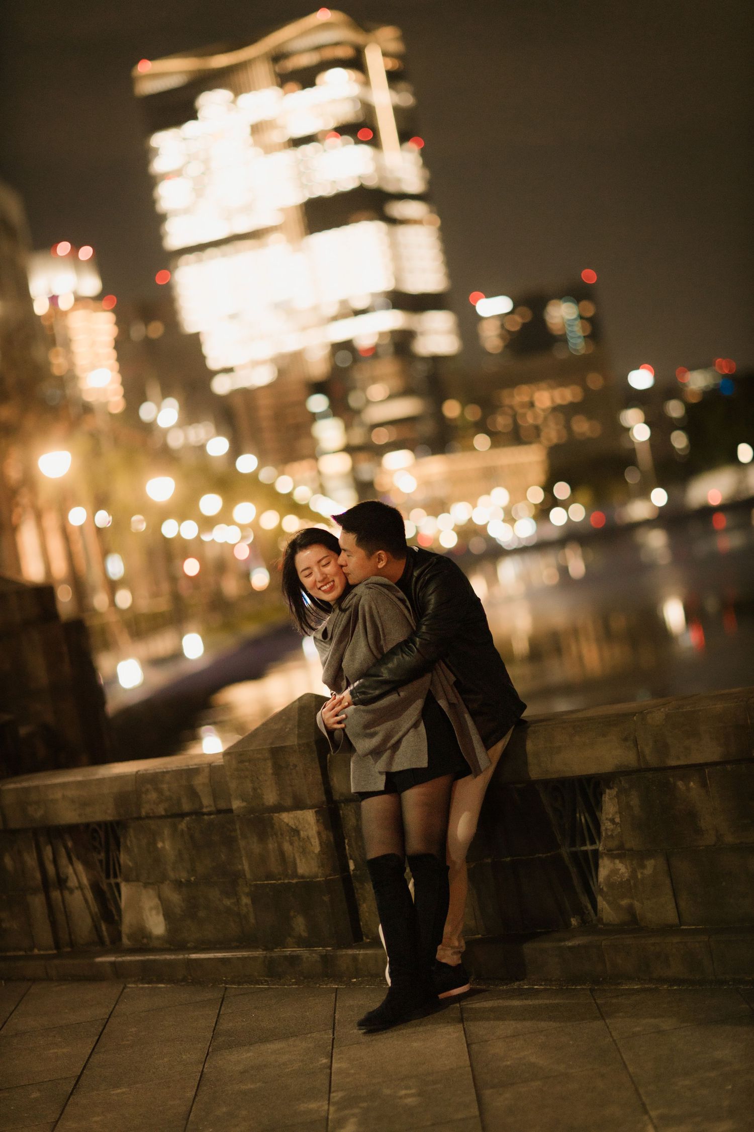 Romantic nighttime cityscape with couple embracing by illuminated buildings.