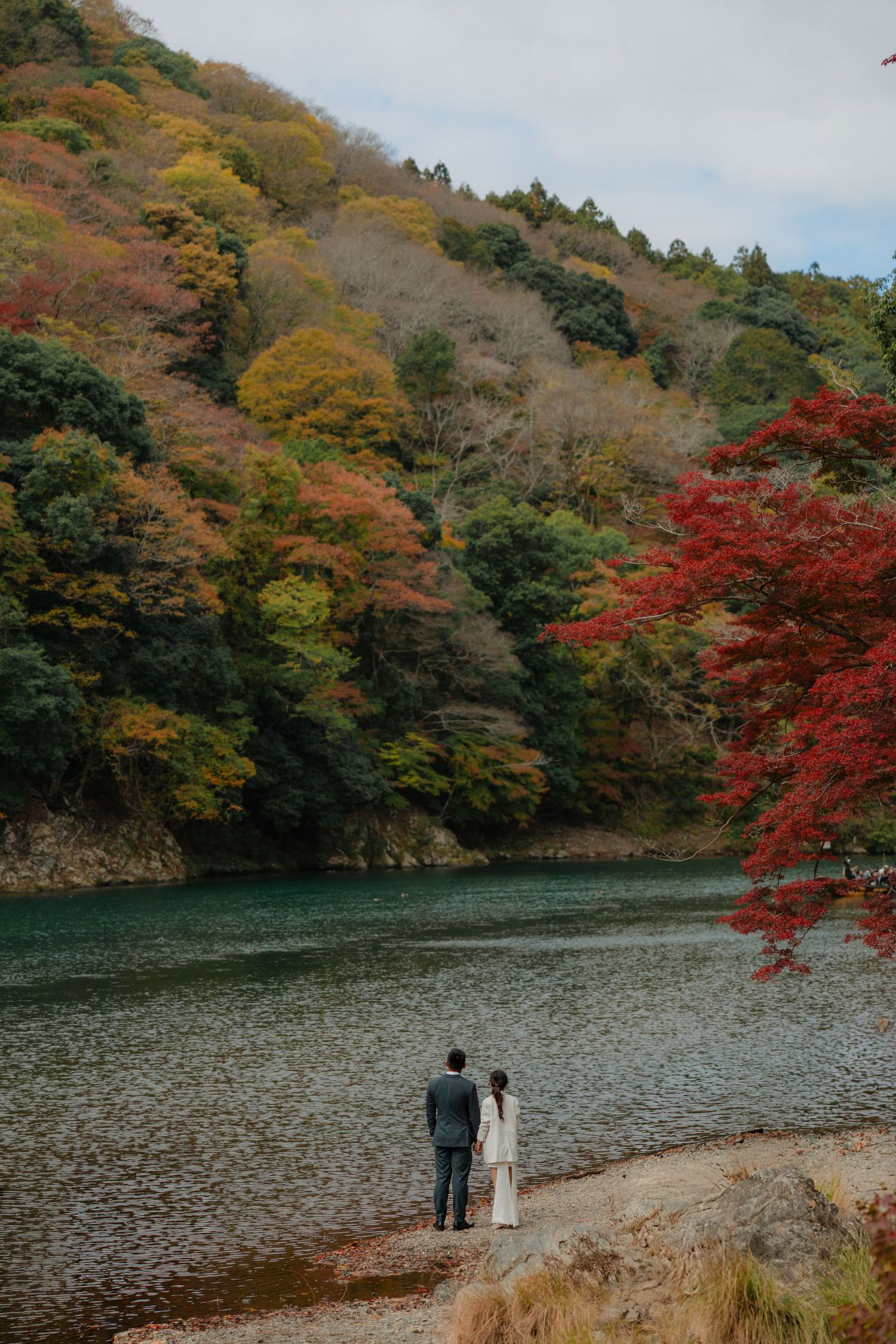 Two people stand at the edge of a lake surrounded by vibrant fall foliage.