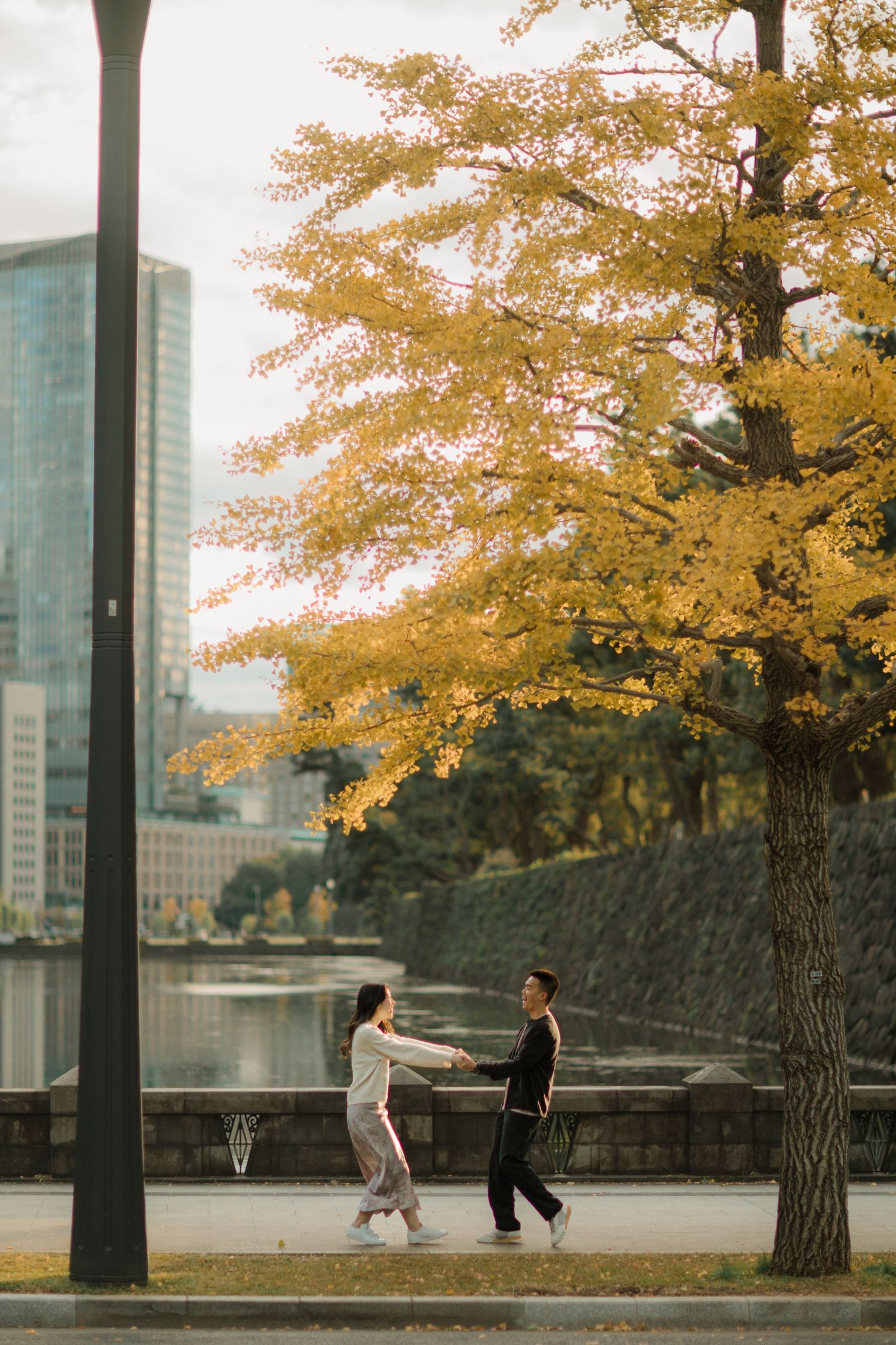 Two people dance playfully beneath golden autumn trees with urban buildings in the background.