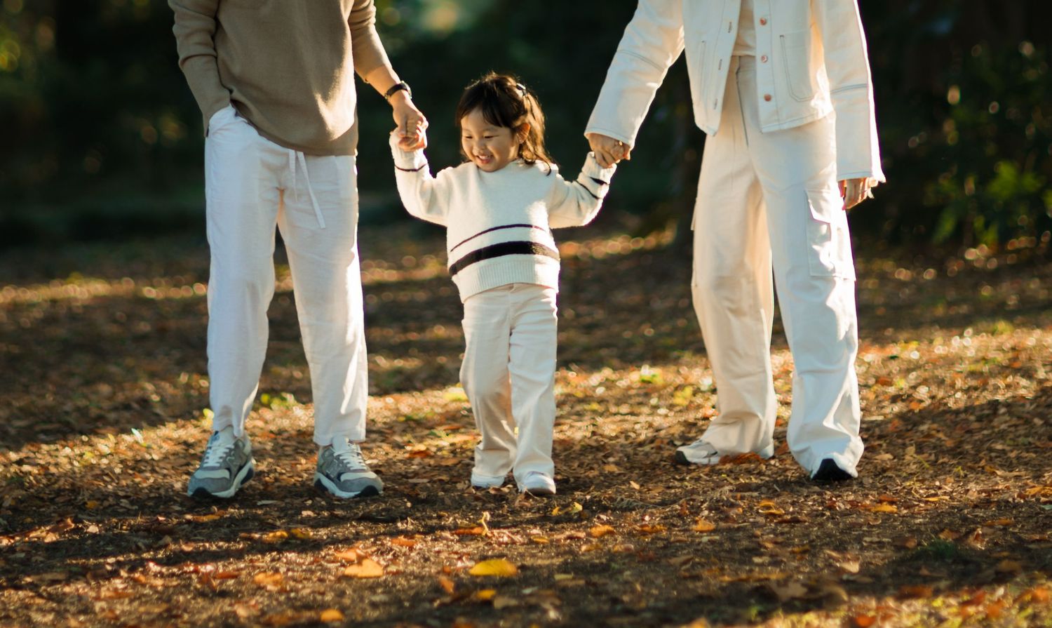 Family members in white clothing hold hands with child during outdoor walk.