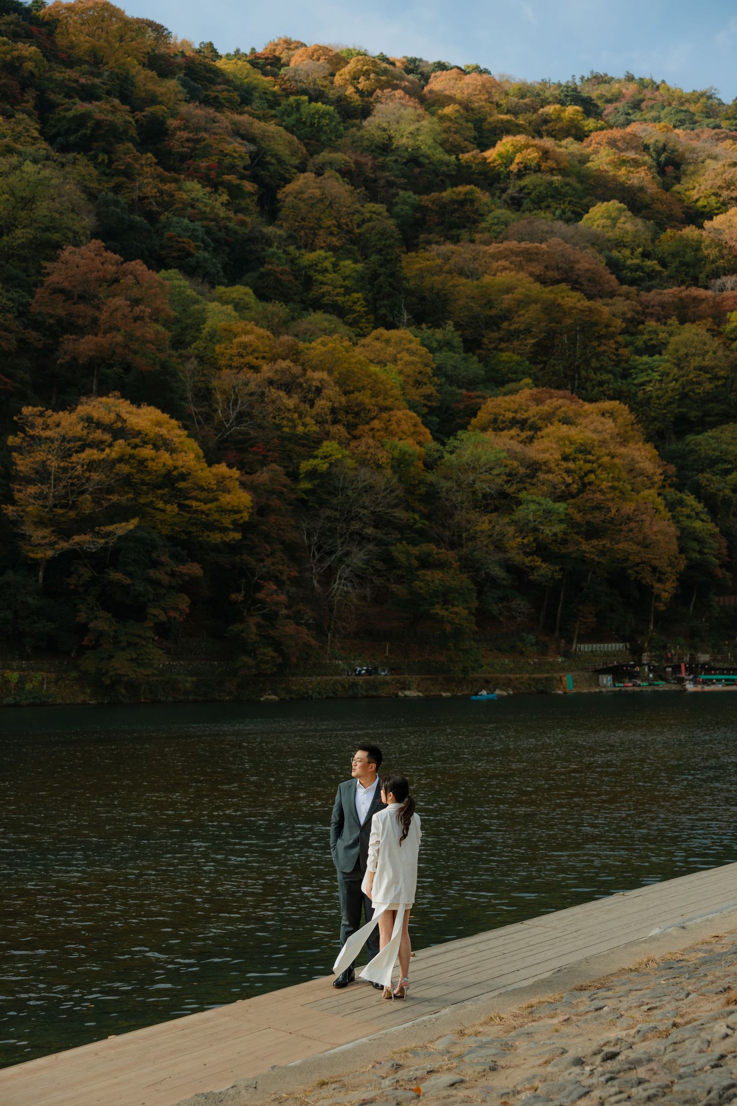 A couple walks along a lakeside path with autumn-colored hills in the background.