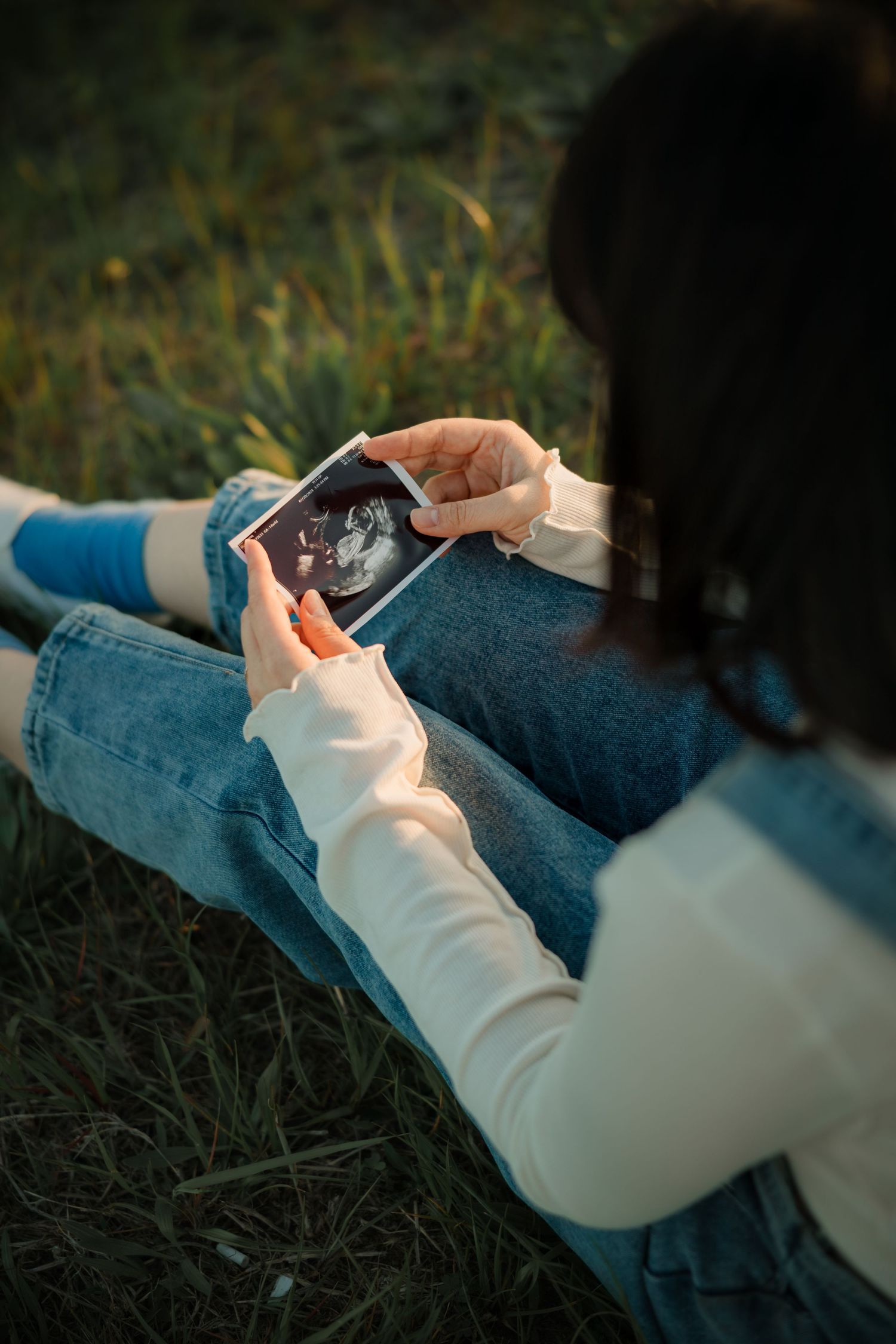 Person holding ultrasound photo while sitting on grass outdoors.