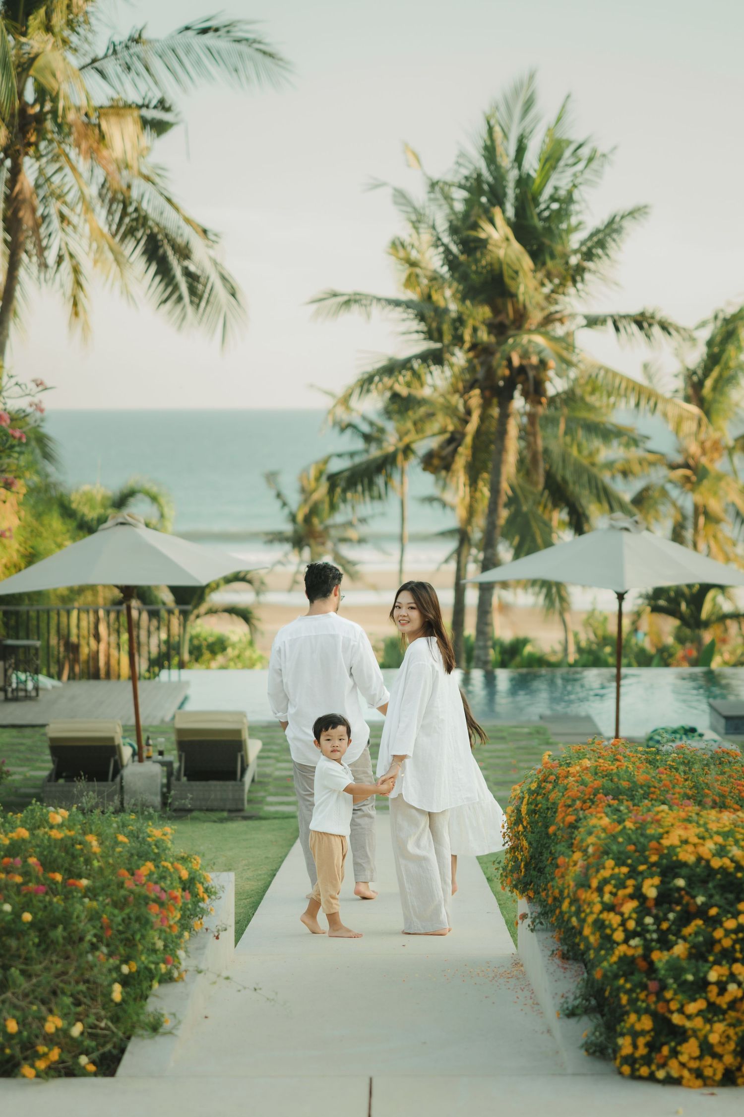 Family walking along tropical resort pathway lined with palm trees and flowers.