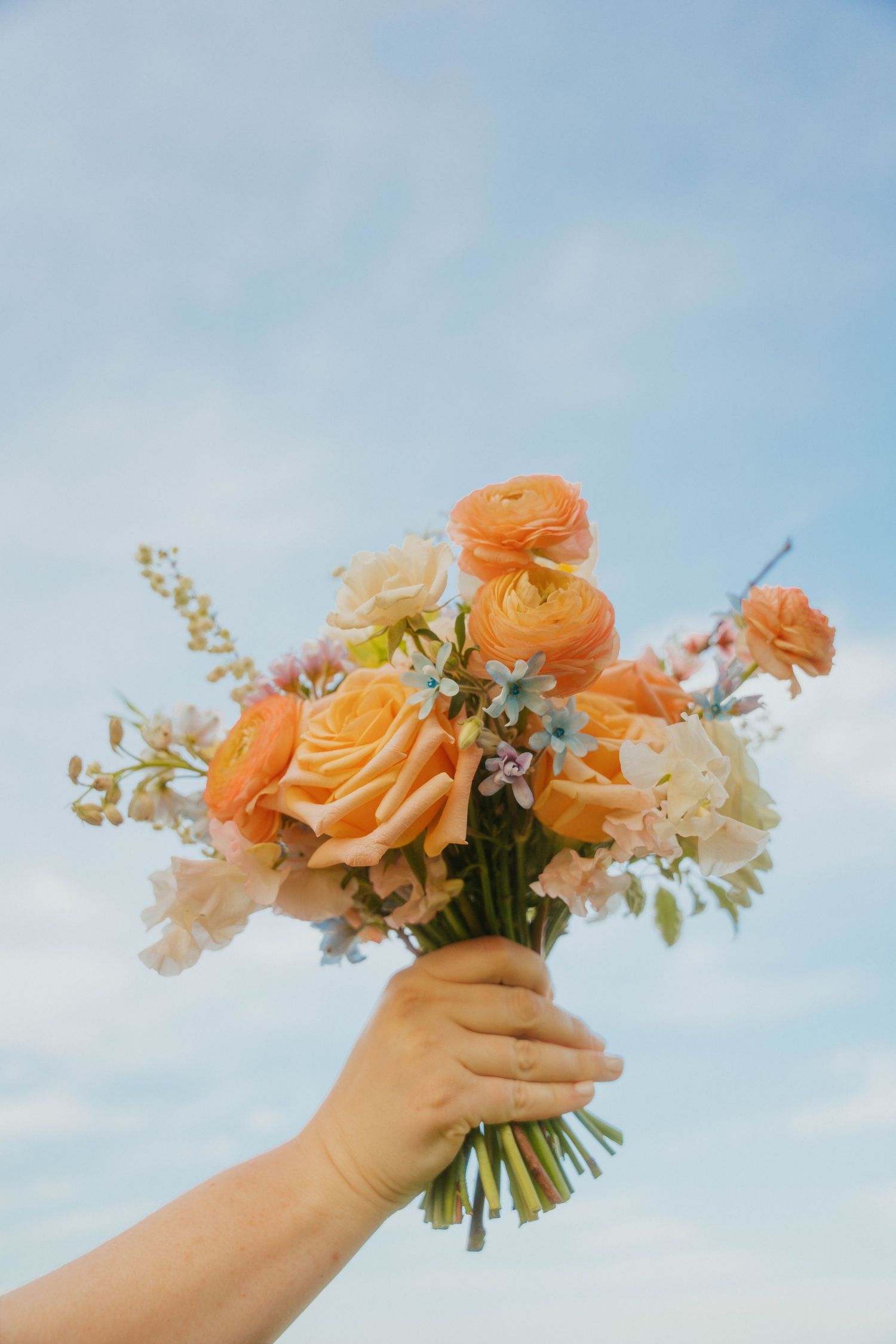 Hand holding a bouquet of peach roses and mixed flowers against a blue sky.