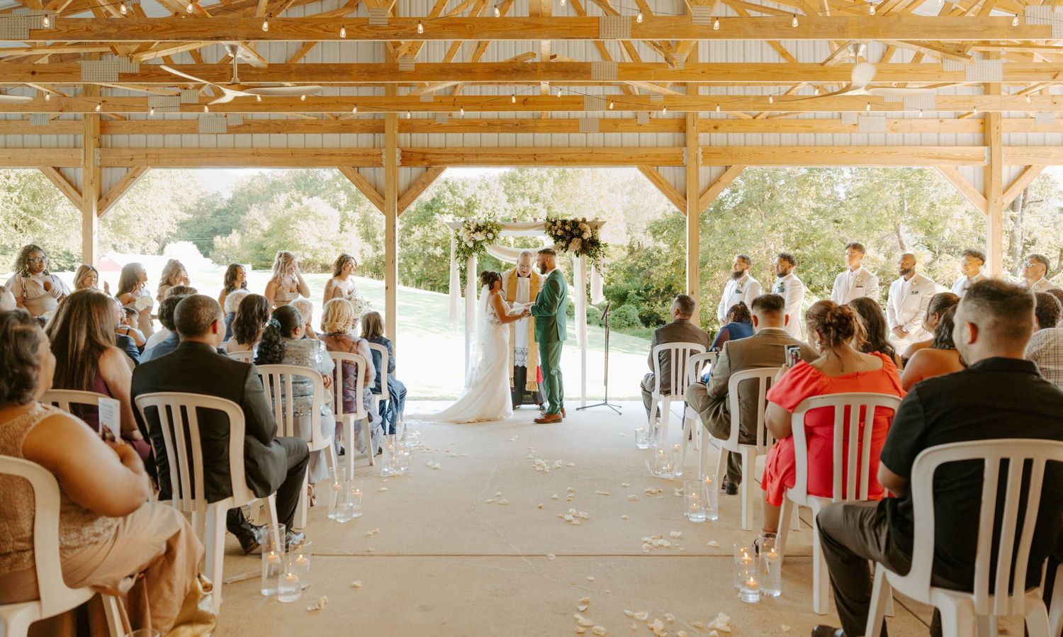 Outdoor wedding ceremony under wooden pavilion with guests seated as couple exchanges vows at altar.