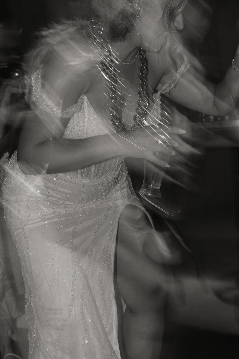 Black and white motion blur photo of a woman in a sparkling white dress and chain necklace dancing energetically.