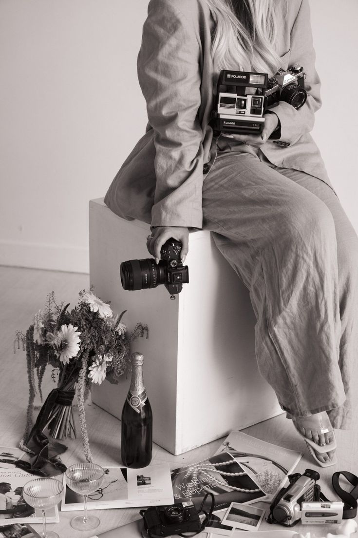Black and white photo of a photographer sitting on a cube holding multiple cameras with flowers and film scattered on the floor.