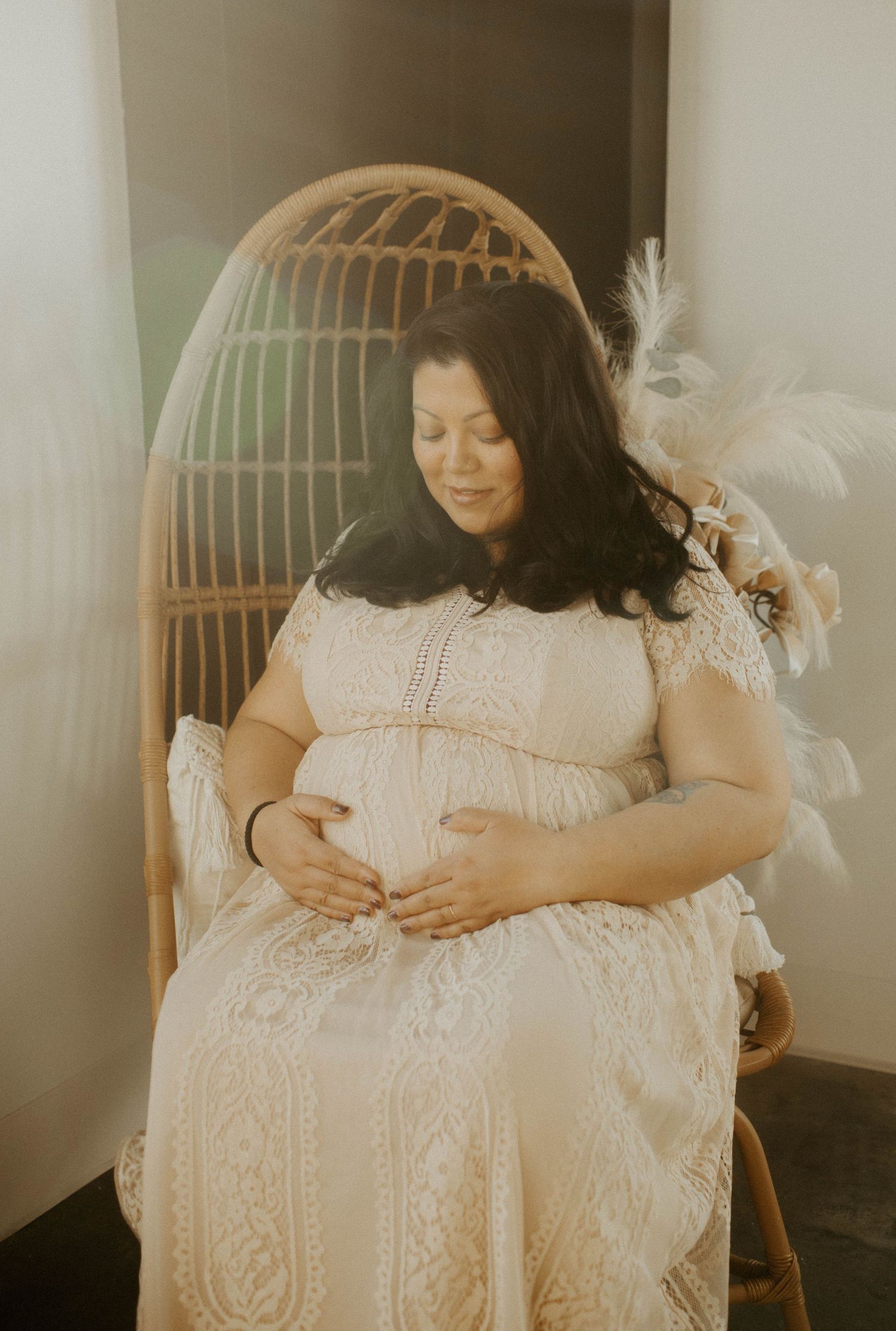 Pregnant woman in a lace dress sitting in a rattan chair holding her baby bump in soft light.