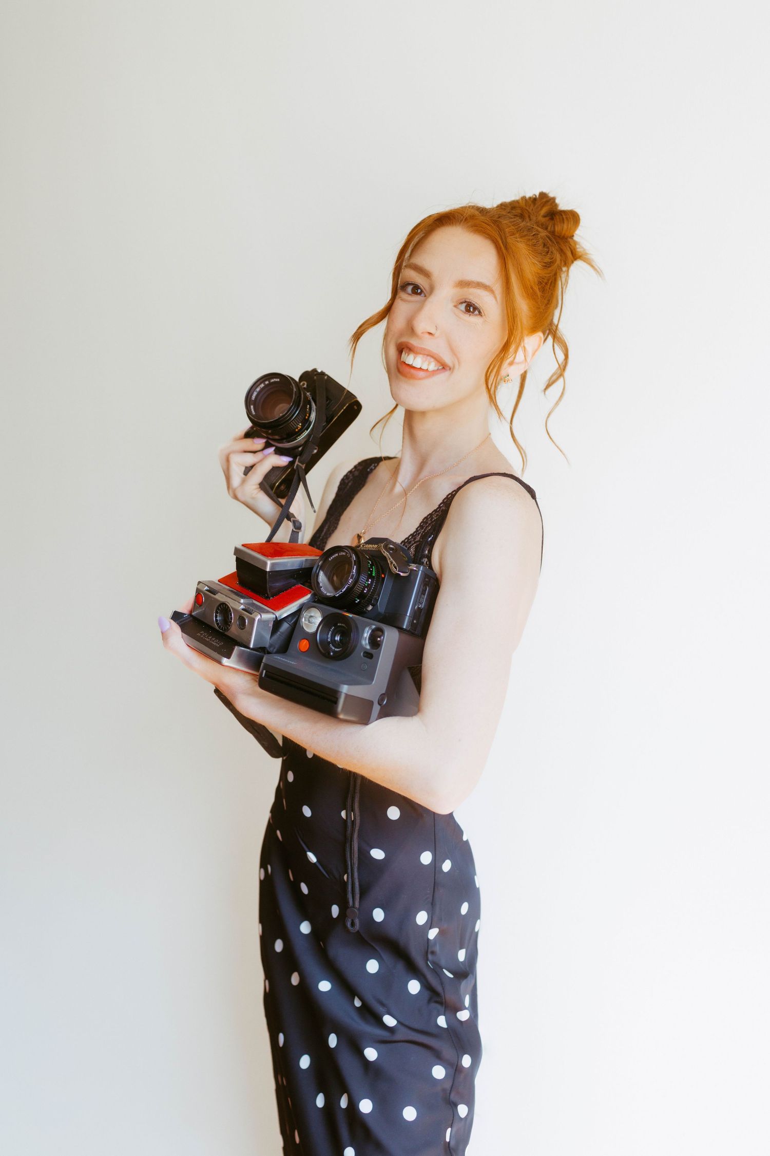 Smiling redhead woman in polka dot dress holding stack of vintage Polaroid and film cameras indoors.