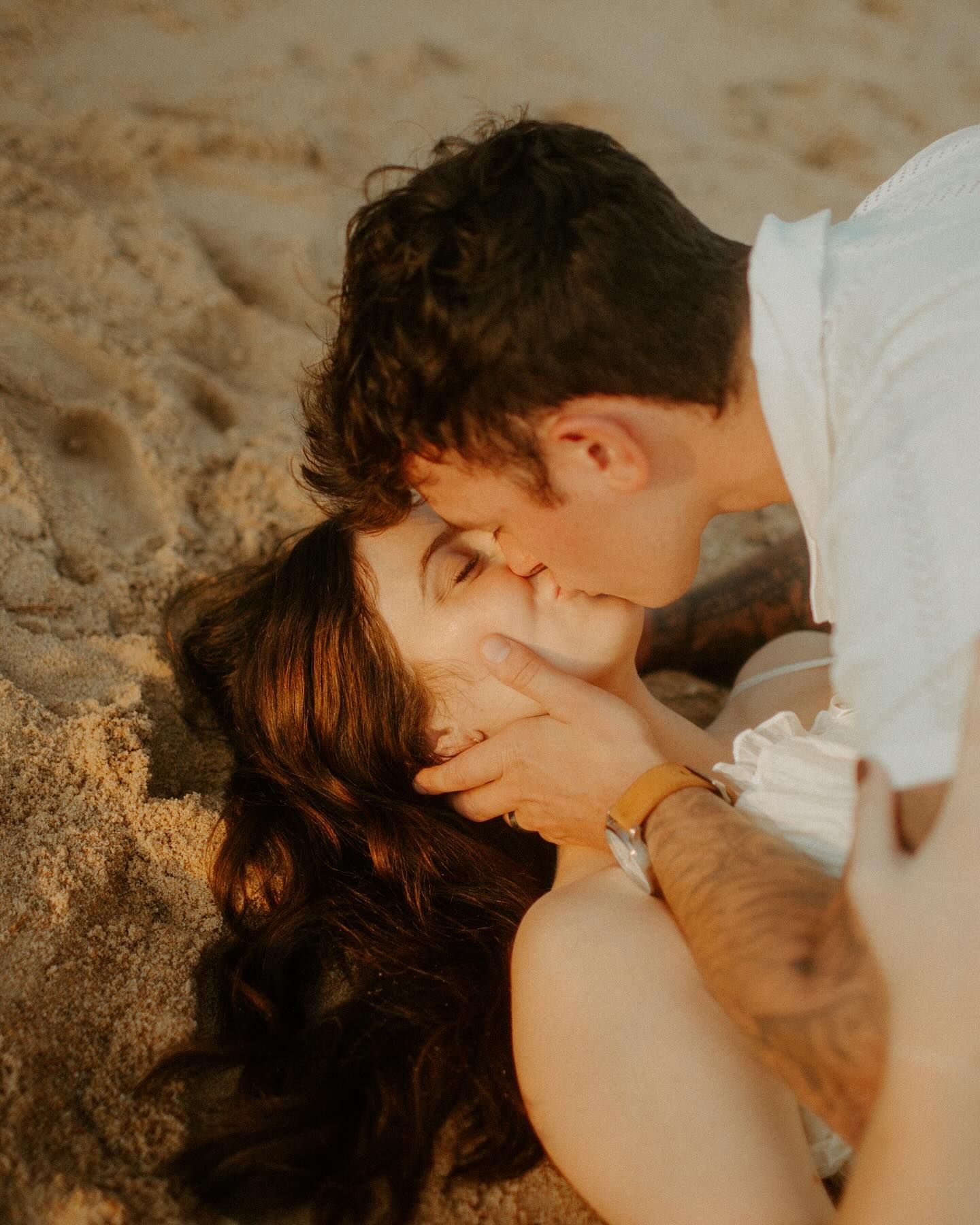 Romantic couple sharing a kiss on sandy beach, man with tattoos tenderly holding woman's face.
