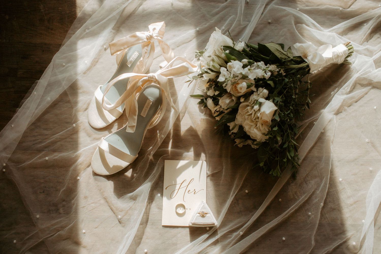 Elegant bridal flat lays with satin heels, white floral bouquet, wedding rings, and veil on sunlit floor.