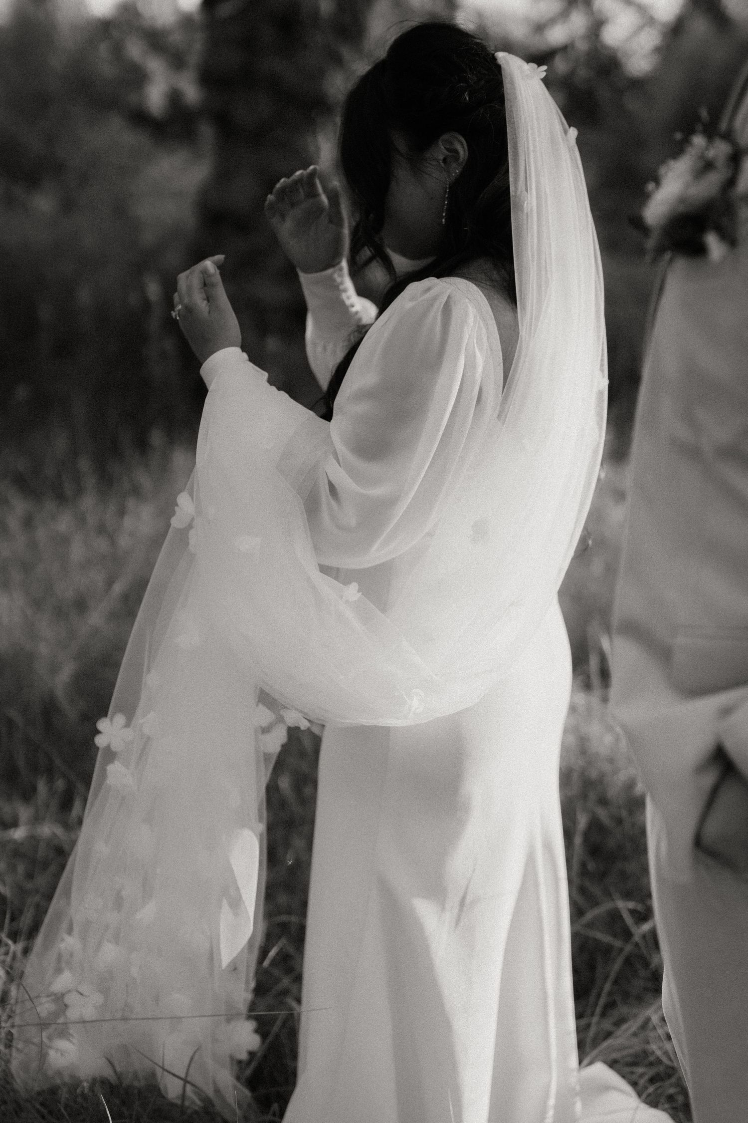 Black and white artistic photo of bride in flowing white gown and floral veil outdoors.