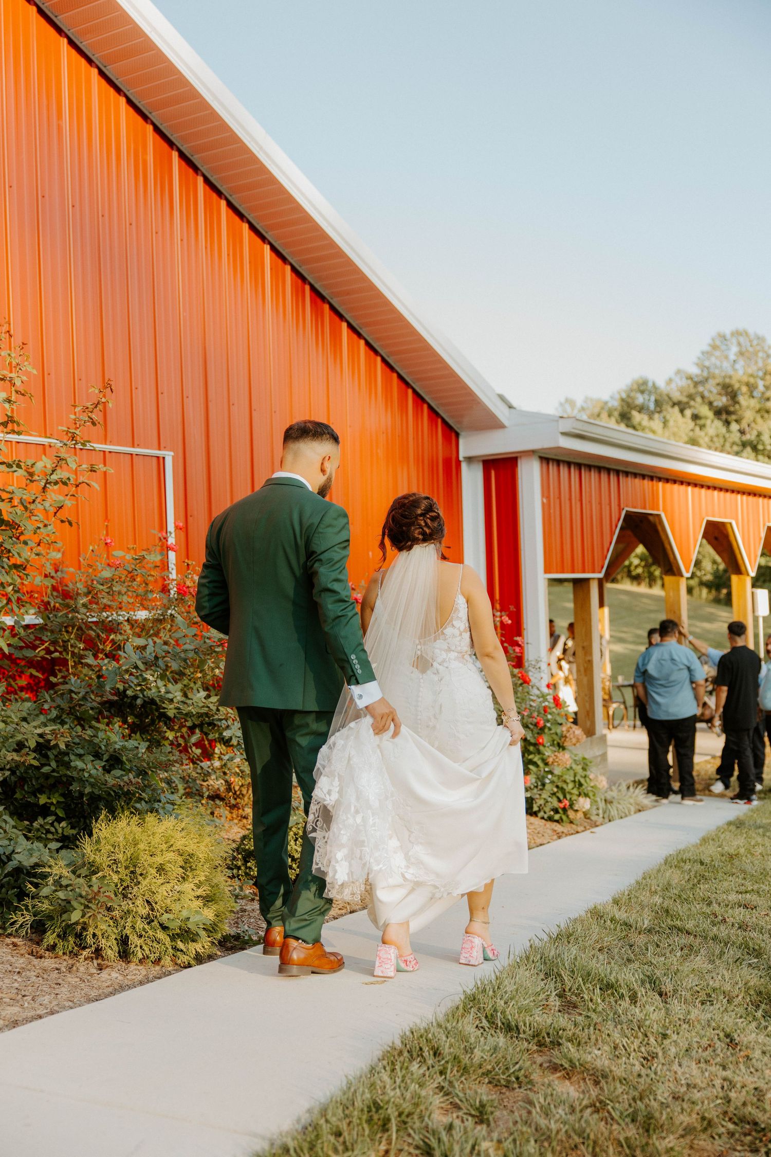 Newlyweds walking hand in hand past a vibrant red barn on their wedding day.