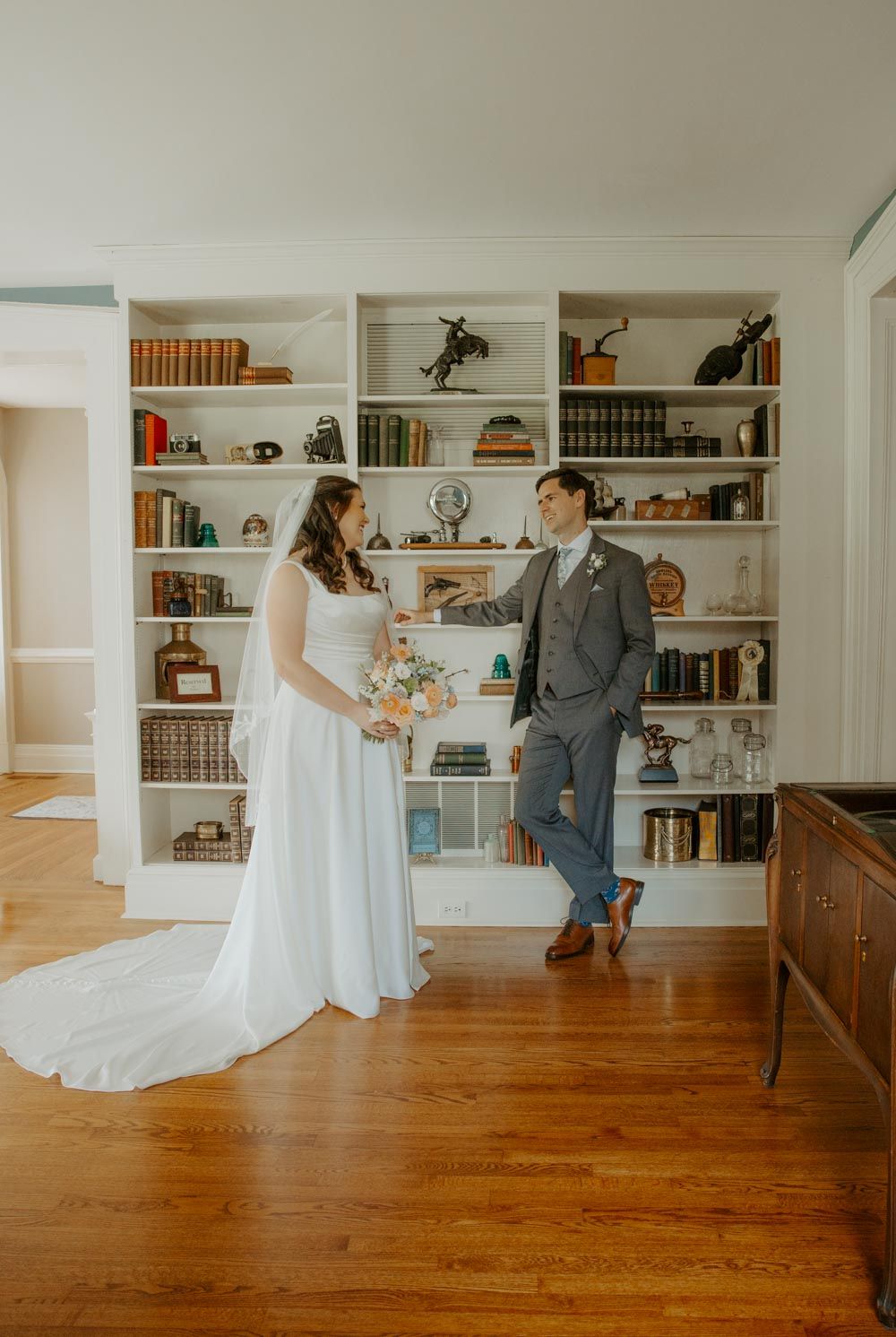 Bride and groom share a tender moment in front of a white bookshelf in an elegant room with hardwood floors.