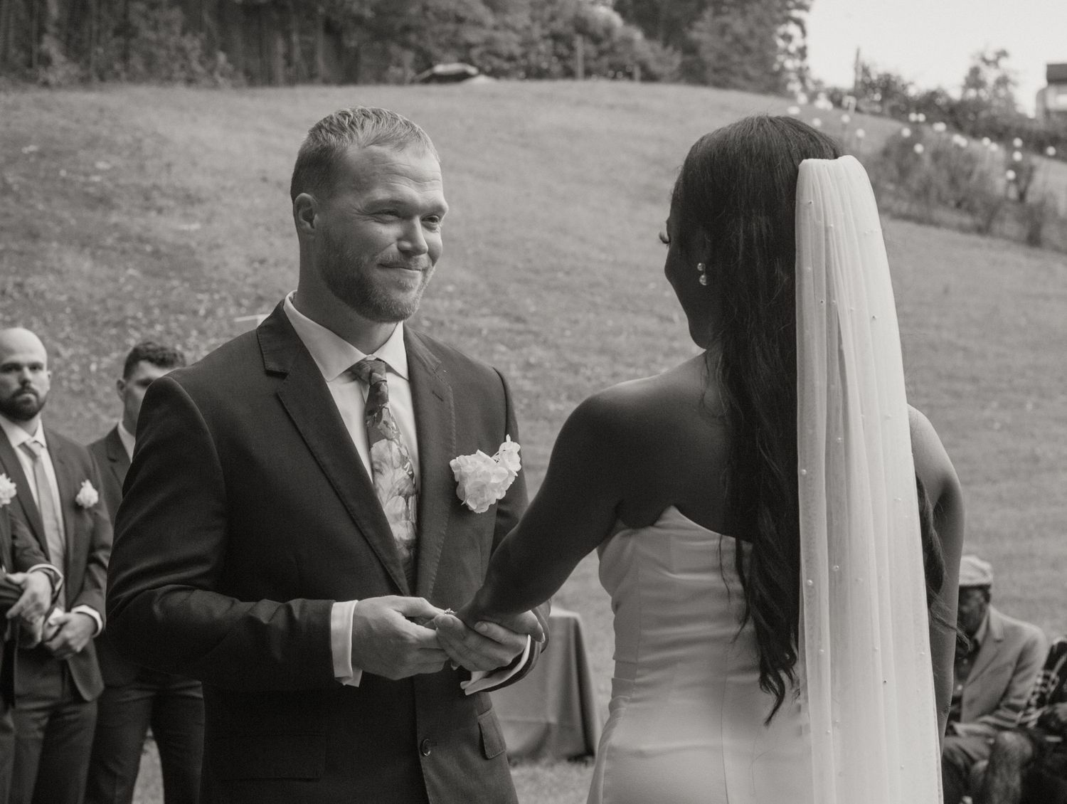 Black and white wedding ceremony photo of smiling groom receiving ring from bride in elegant veil outdoors.
