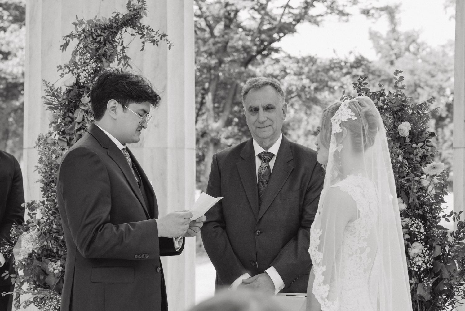 Groom reads vows to veiled bride at outdoor wedding ceremony while officiant watches in black and white photo.