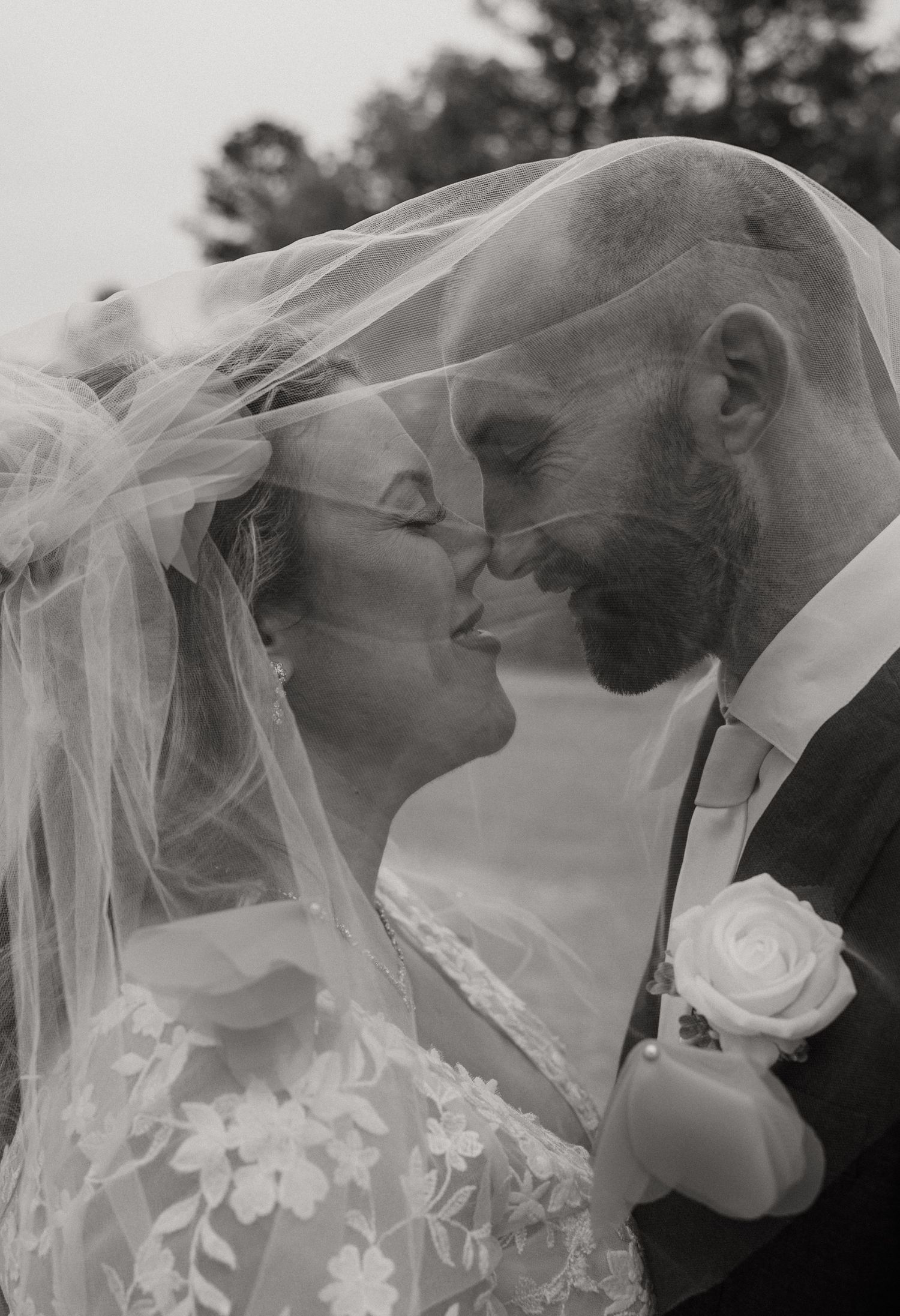 Black and white wedding photo of bride and groom sharing a kiss under a flowing veil, bride in lace gown.