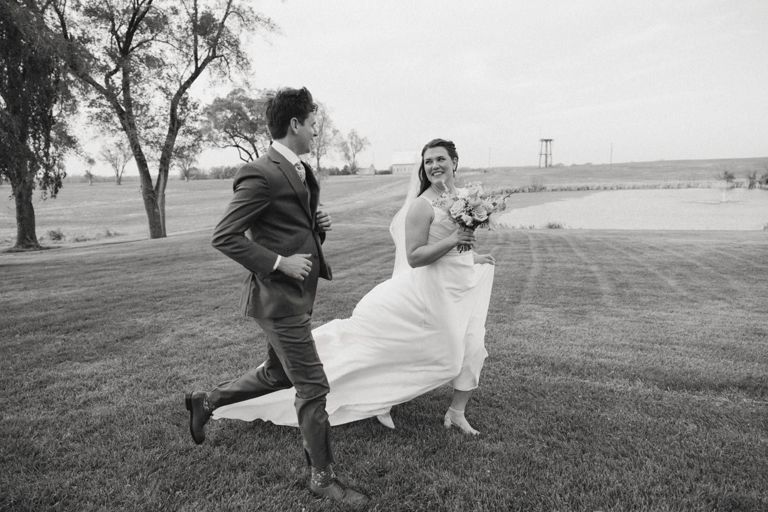 Black and white photo of a bride and groom playfully running on a grassy lawn during their wedding day.