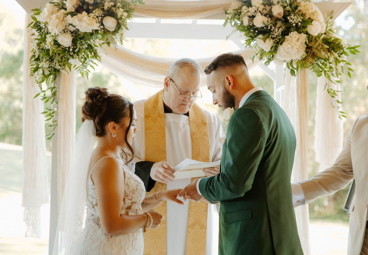 Bride and bearded groom exchange vows under floral arch with officiant at outdoor wedding ceremony.
