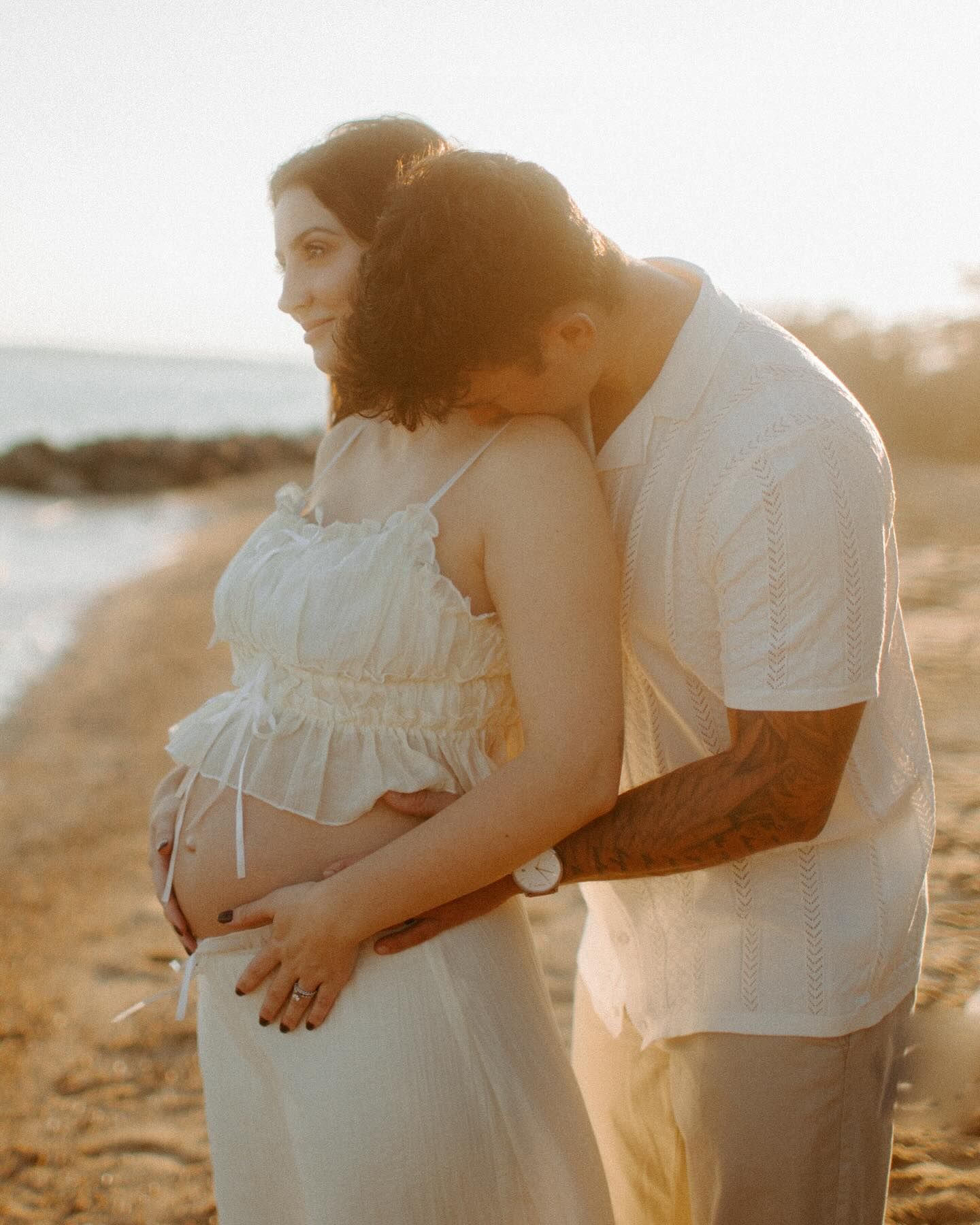 Couple tenderly embracing on a sandy beach at golden sunset, hands cradling a pregnant belly.