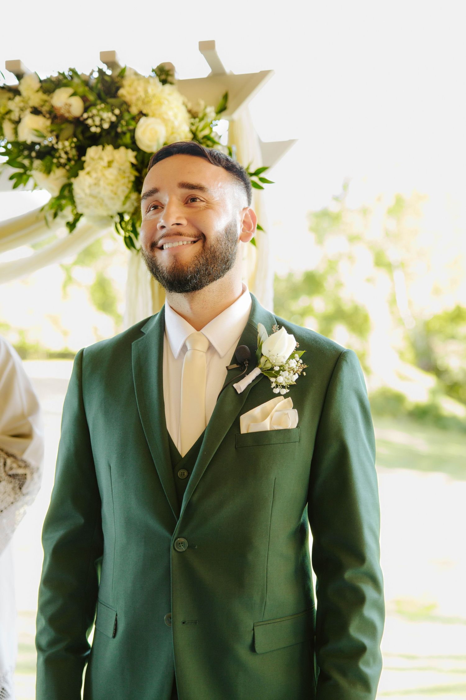 Smiling groom in a green three-piece suit with white boutonniere at outdoor wedding altar.