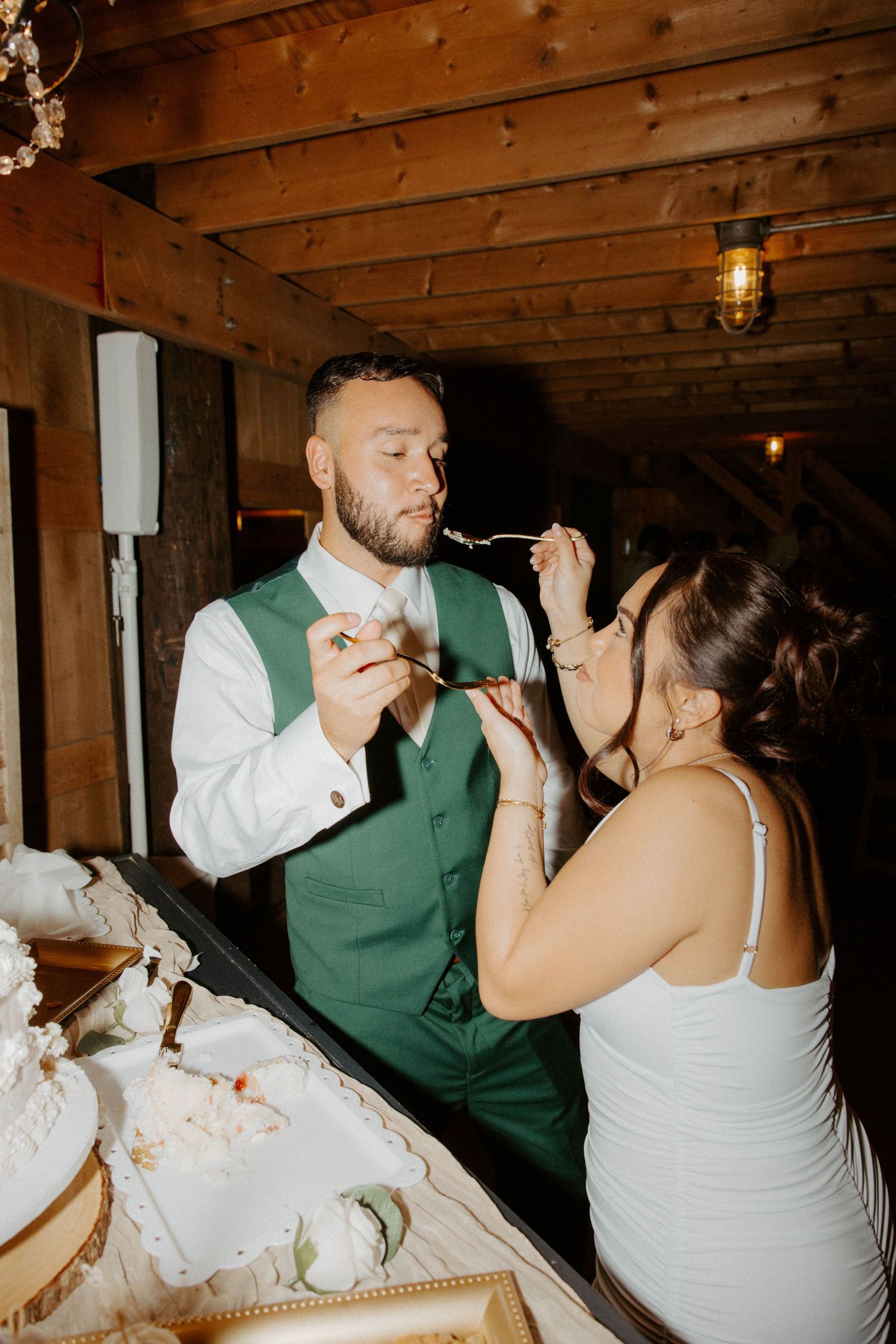 Bride and groom feeding each other wedding cake at rustic barn reception.